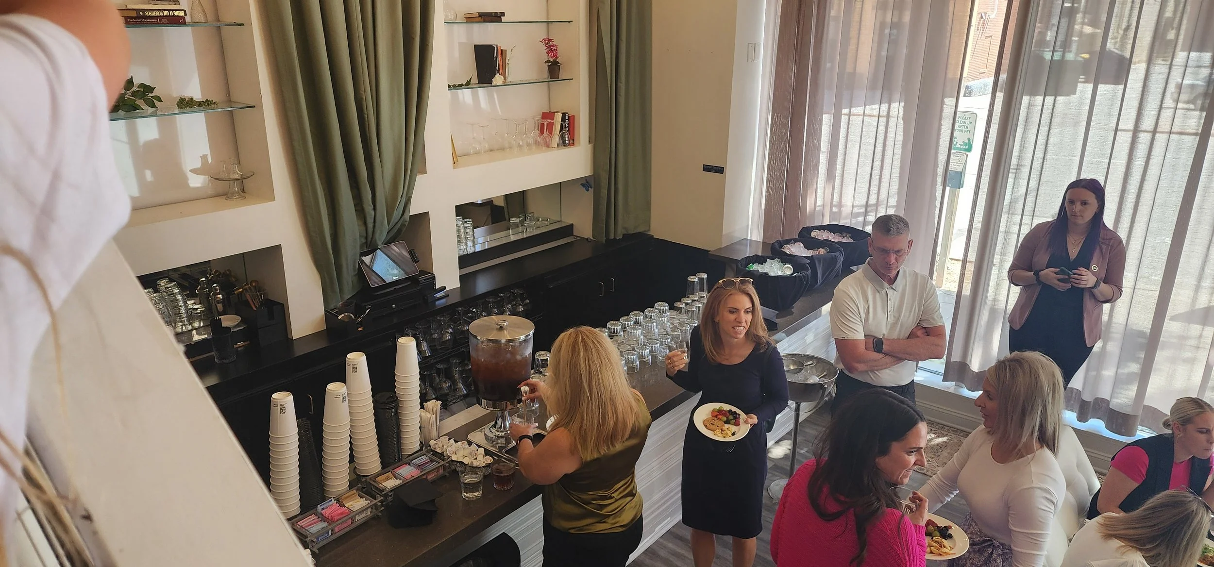 Group of people socializing in a room with a refreshment table, glasses, stacked cups, and coffee dispensers. Some individuals are holding plates with food.