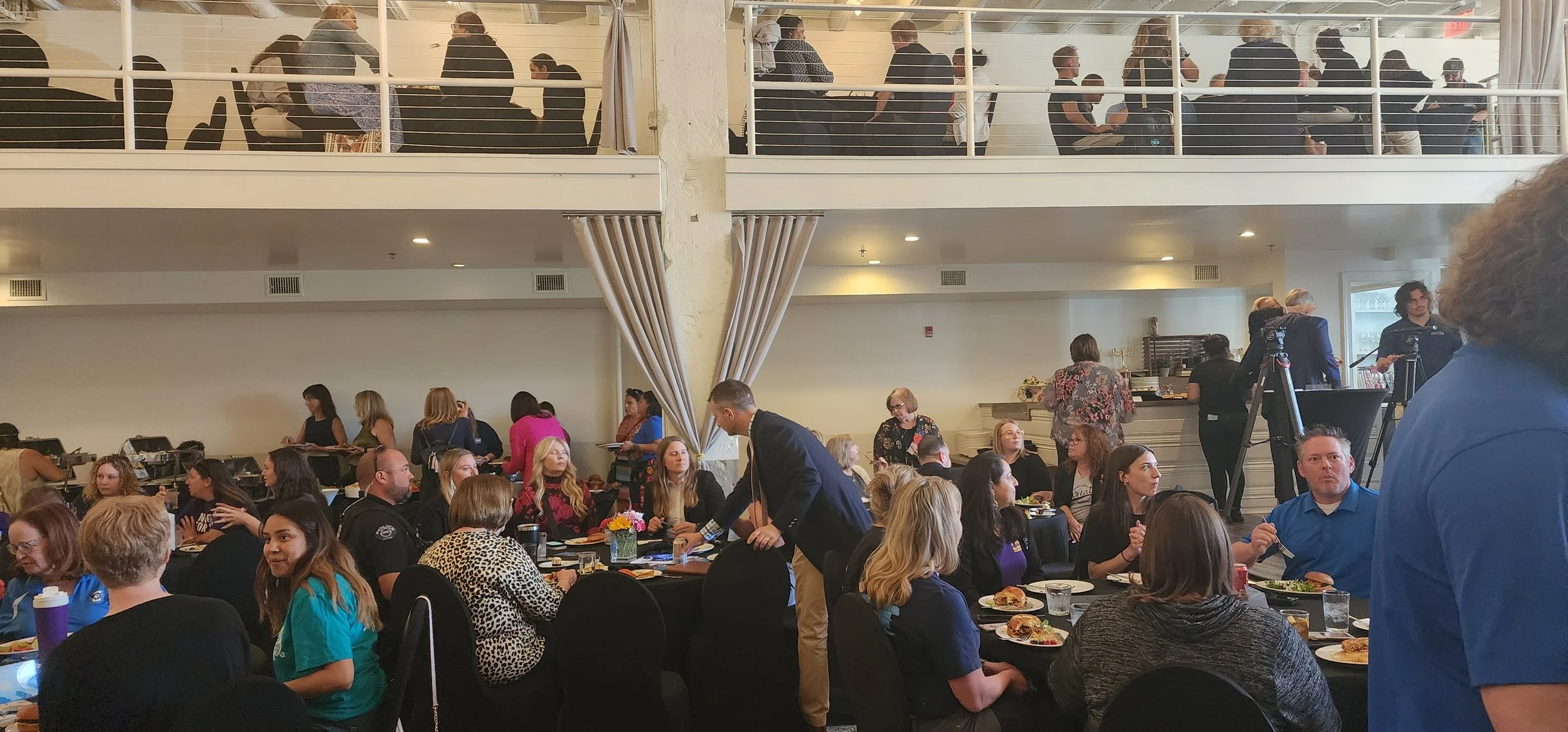 People gathered at an indoor event, seated at tables with food, and others standing. There's an upper level with more people sitting.
