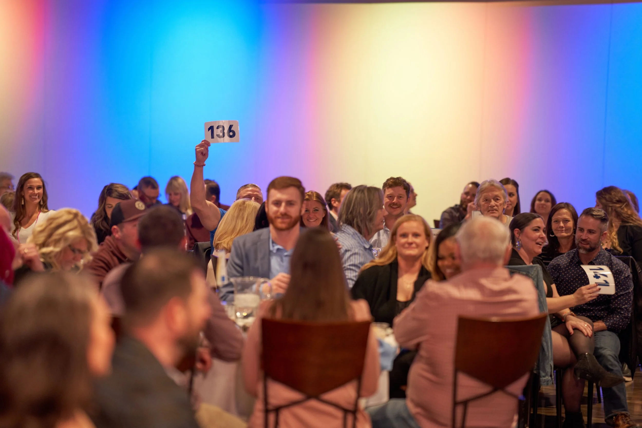 People seated at tables during a public auction, one person holding a bid card with the number 136 visible, colorful lighting on walls.