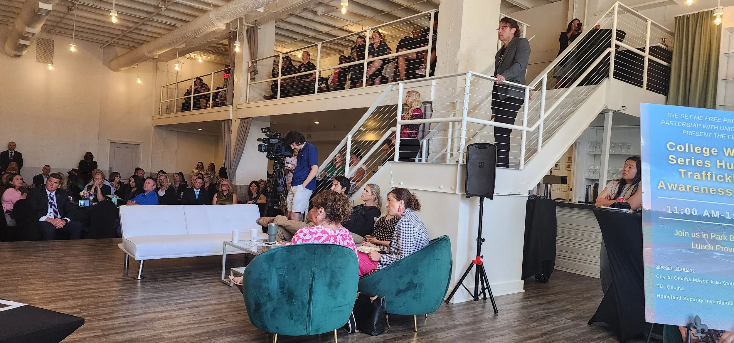 Event at a modern venue with attendees seated and standing, a photographer capturing the scene, and a poster about a human trafficking awareness event. The space includes both main floor and balcony seating with minimalist decor.