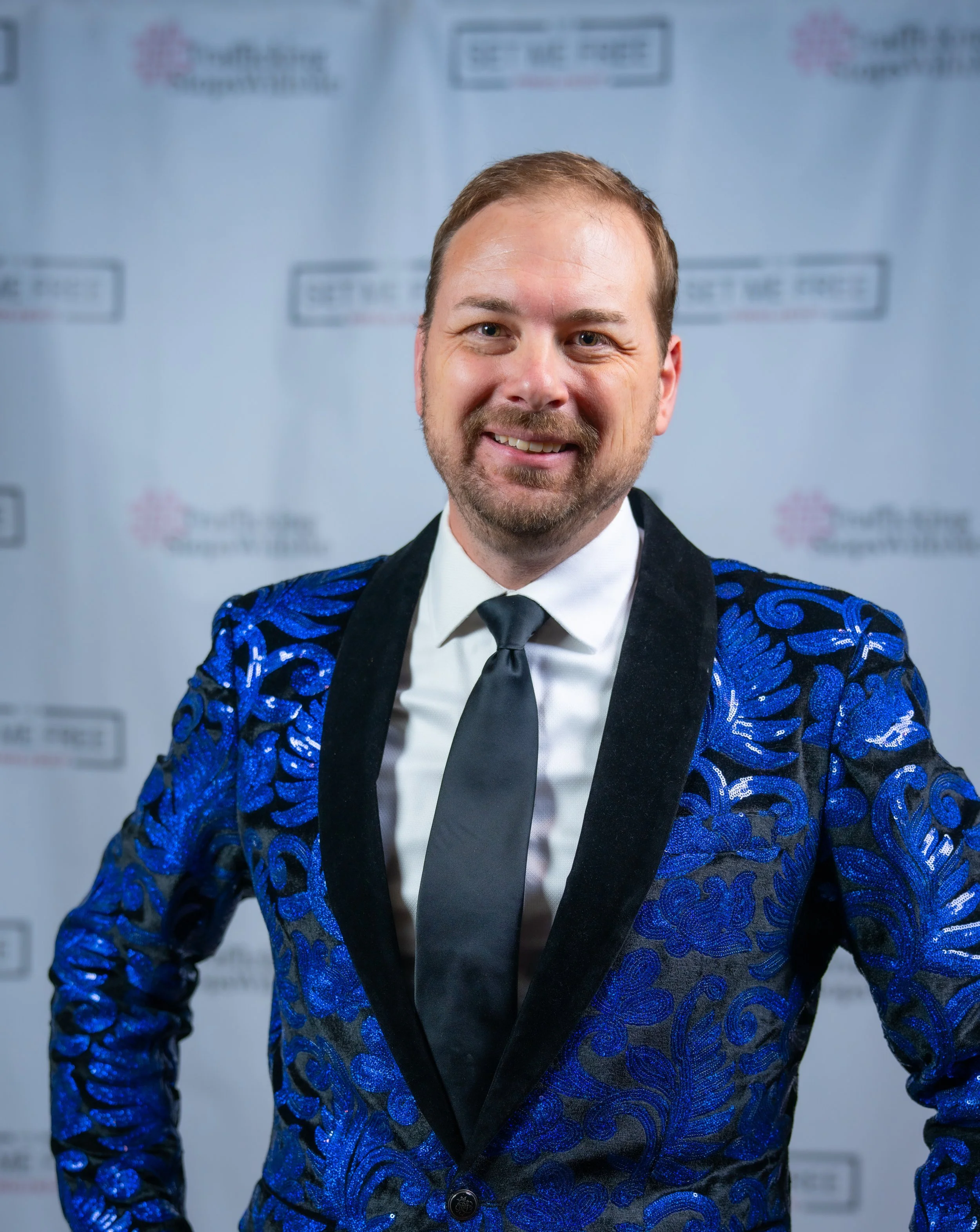 Man in a blue floral jacket with a black tie standing against a backdrop.