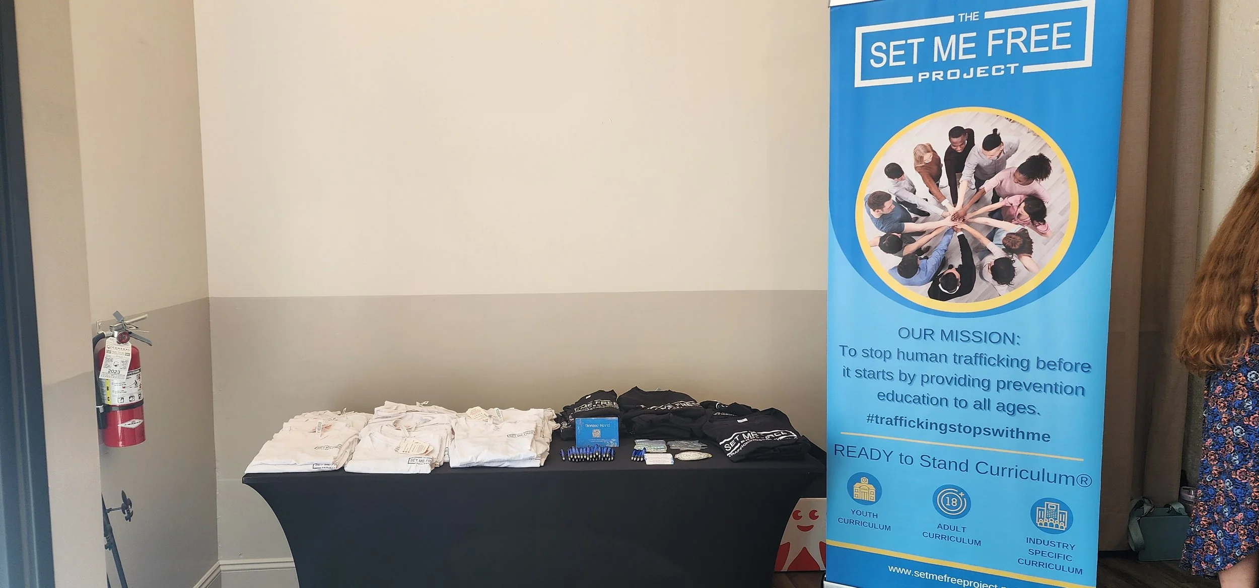 Table with informational banner for 'Set Me Free Project', displaying t-shirts and promotional materials. The banner discusses human trafficking prevention and educational programs.