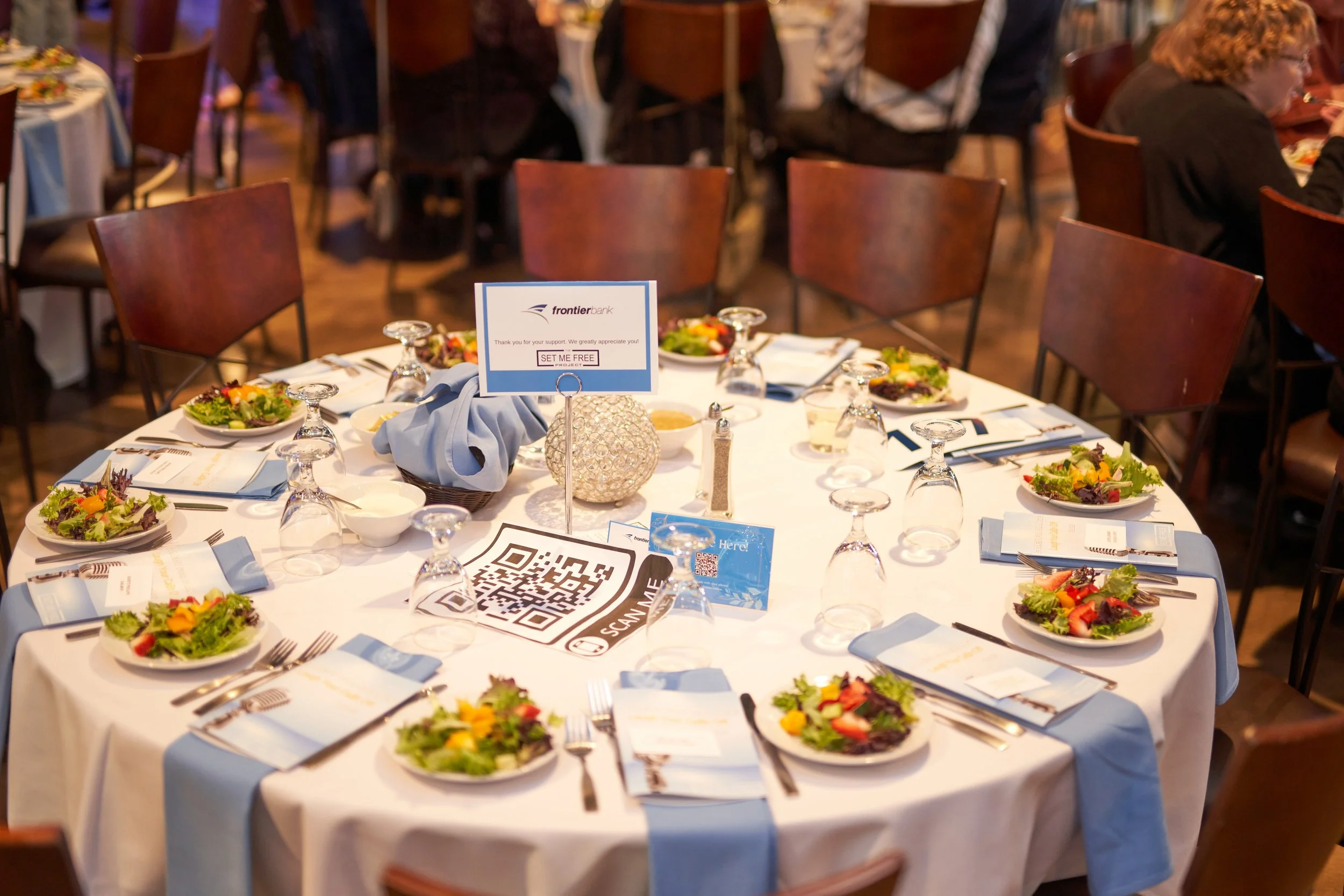 A formal dining table set for an event with salads and place settings for eight. The table has blue napkins, a centerpiece, and a table card displaying "Frontier" alongside a QR code. Other tables and guests are visible in the background.