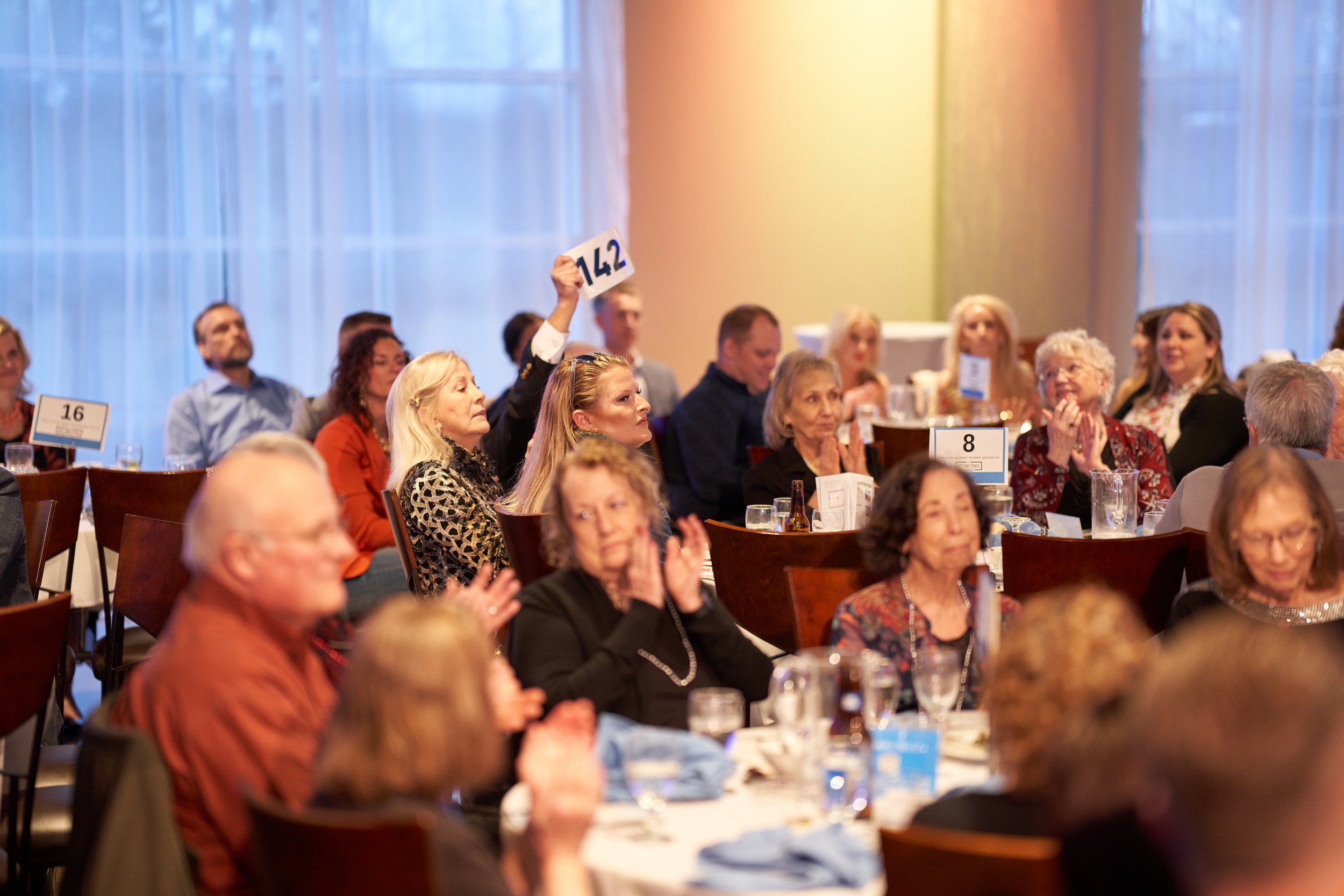 A group of people seated at tables during an auction event. One person is holding up a bid card numbered 142. Attendees appear engaged, with some clapping. Table numbers 8 and 16 are visible. The setting suggests a formal or fundraising event.