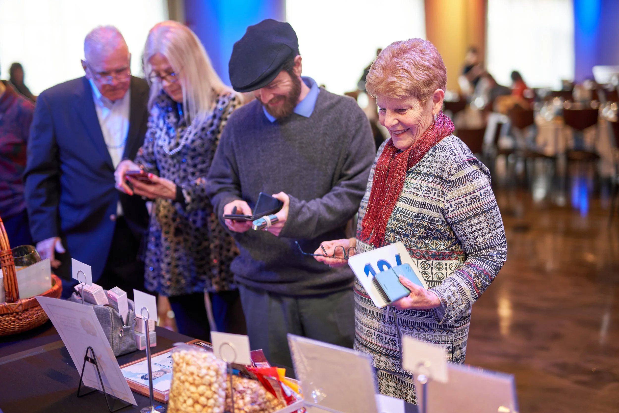 People gathered around a table at an event, holding phones and looking at items on display, possibly for auction.