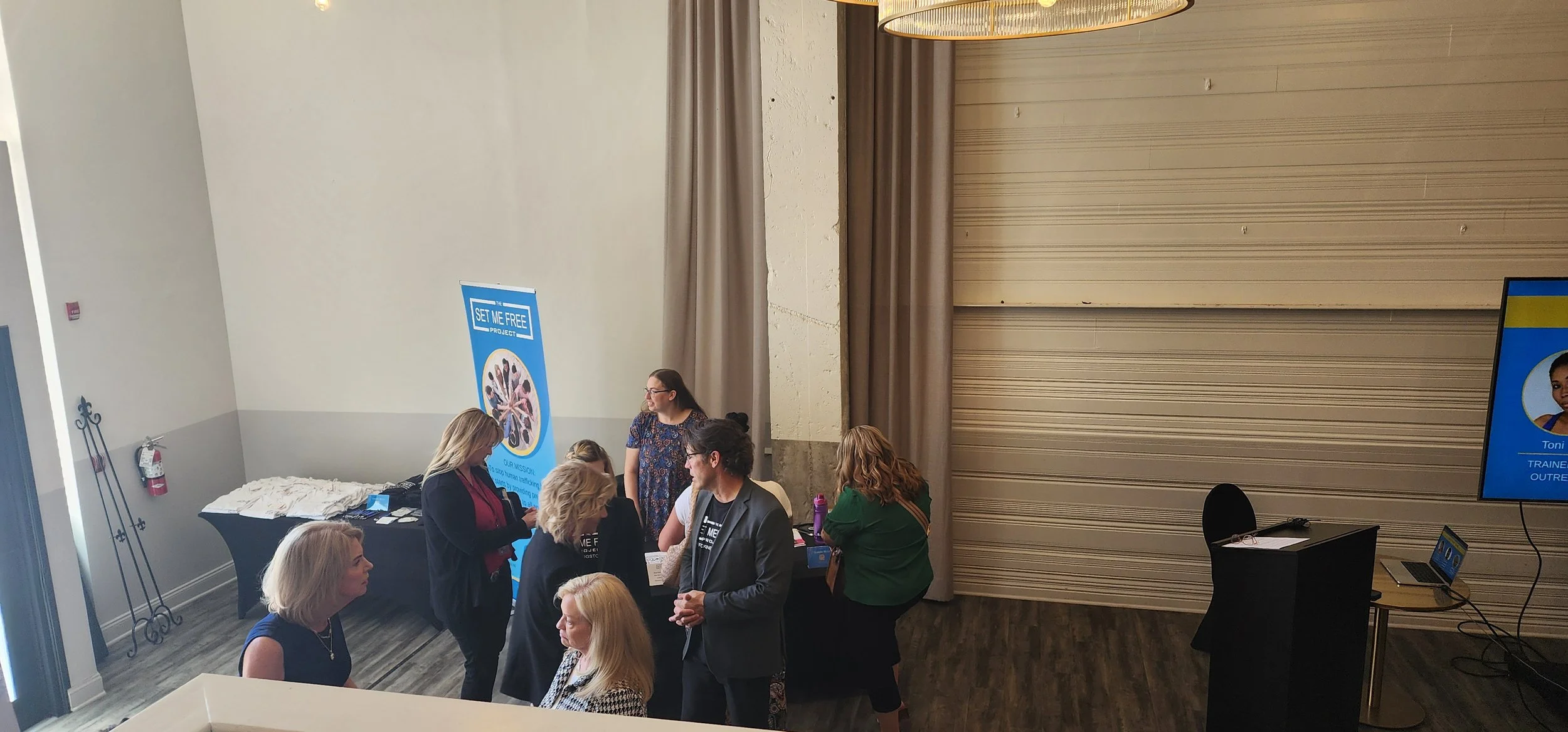 A group of people at an indoor event near a "Set Me Free Project" banner next to tables with promotional materials and a TV screen displaying a name.