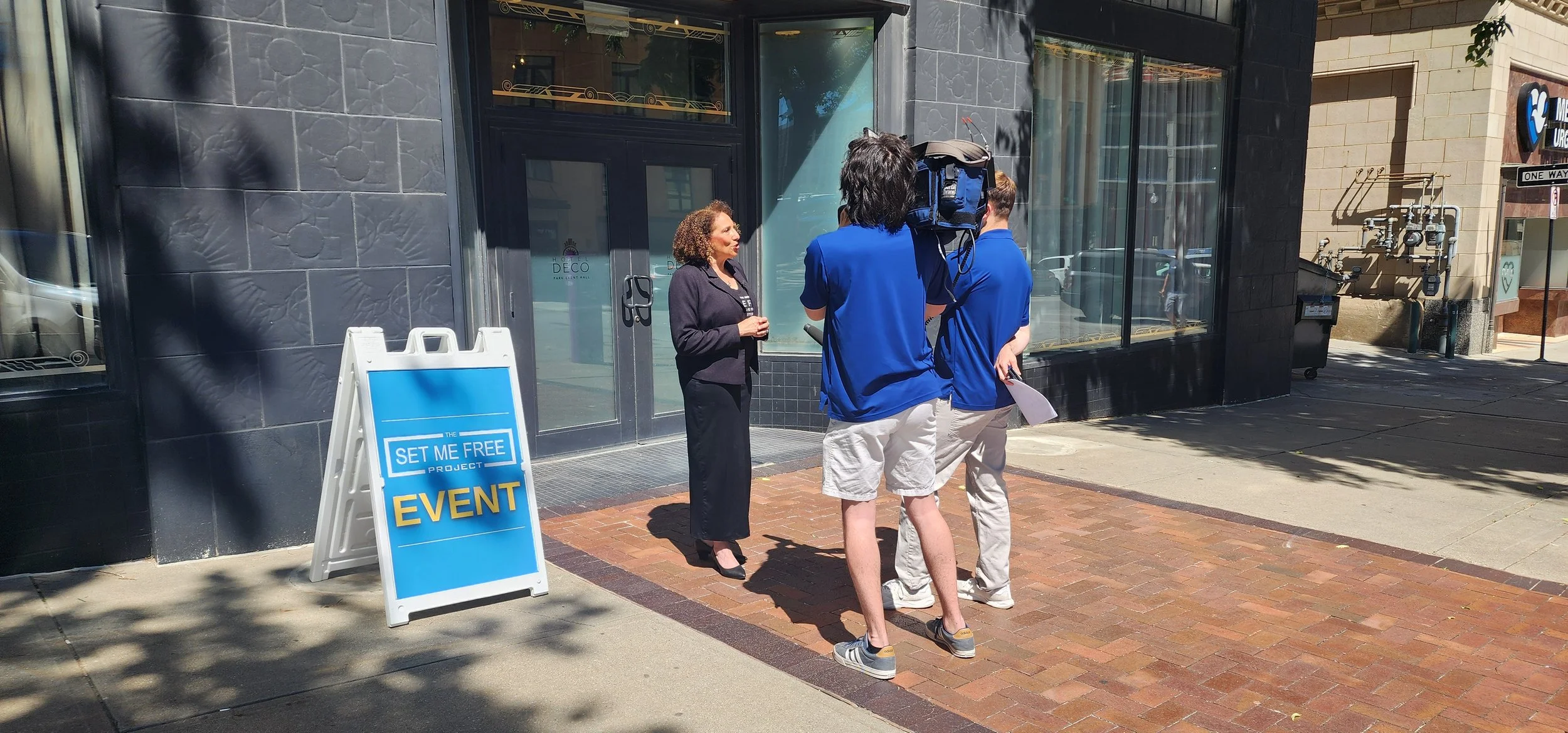 A woman being interviewed by two individuals with a camera outside a building. A sign reads 'Set Me Free Project Event.' The location has dark brick walls and large glass windows.