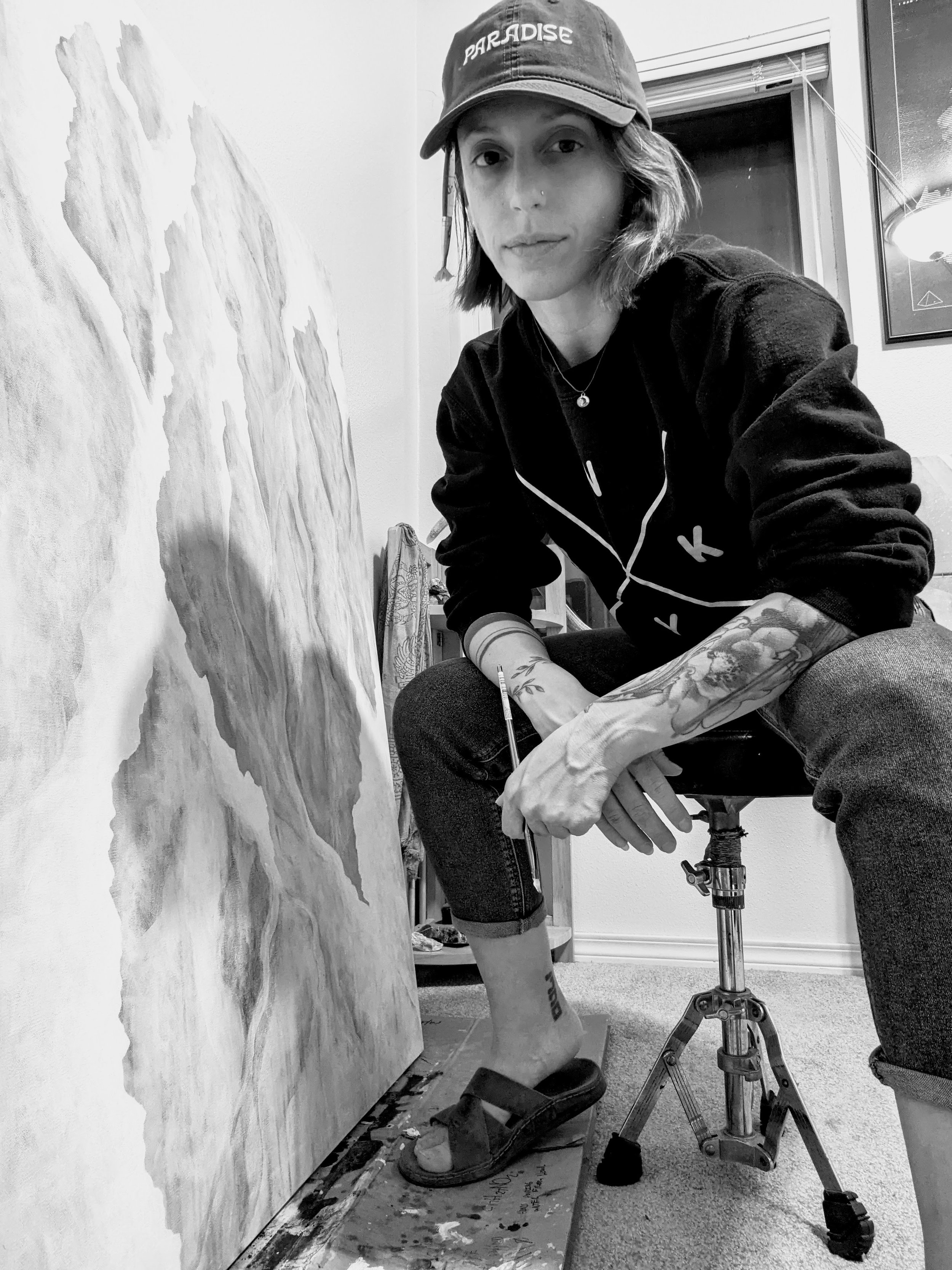 Black and white photo of an artist sitting on a stool, holding a paintbrush, next to a large canvas in a studio. The artist is wearing a cap and casual clothes with visible tattoos.