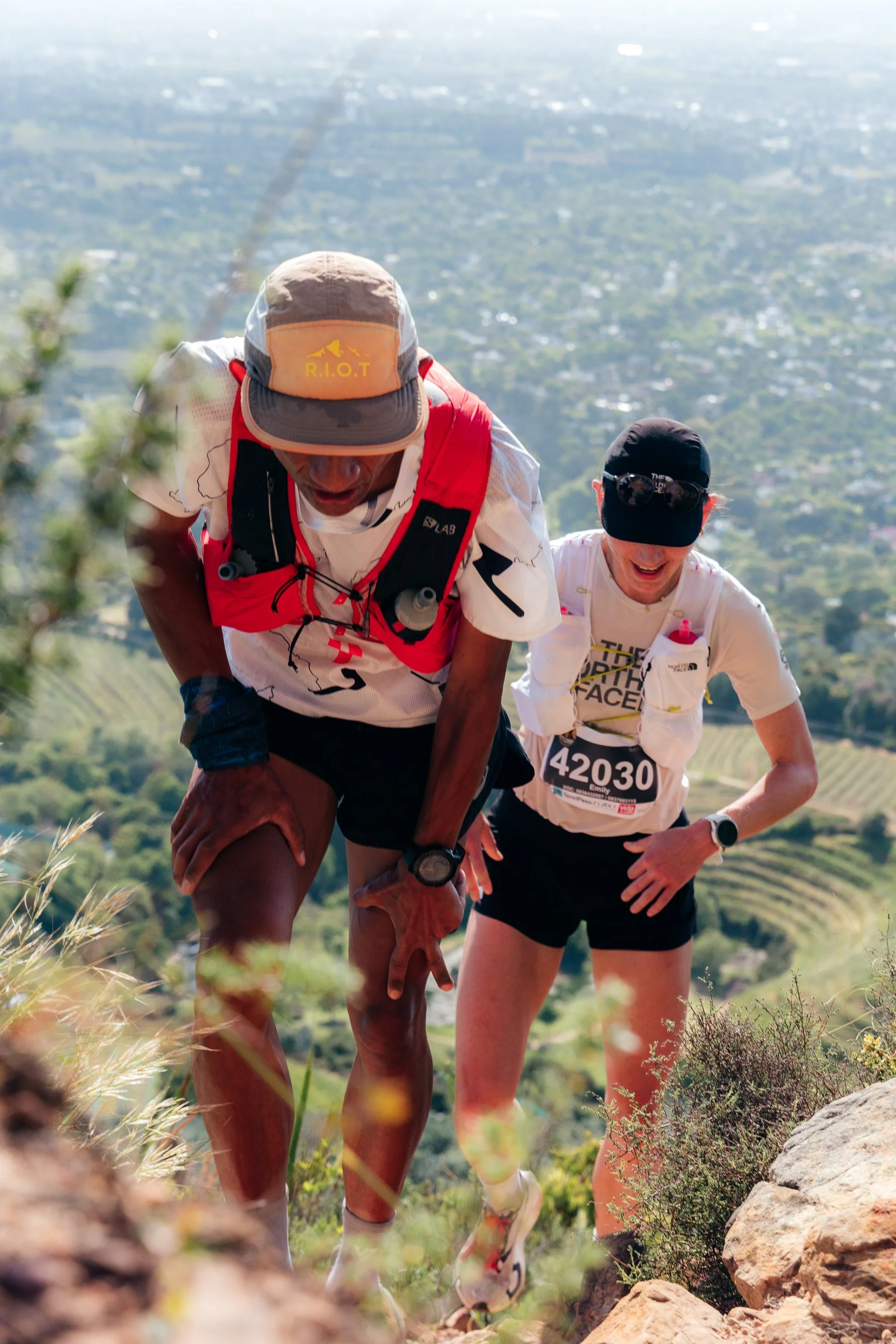 Two trail runners, one wearing a beige cap with 'R.I.O.T' and a red hydration pack, and the other wearing a black cap and sunglasses, climb up a rocky trail with a scenic landscape of green hills and a city in the background.
