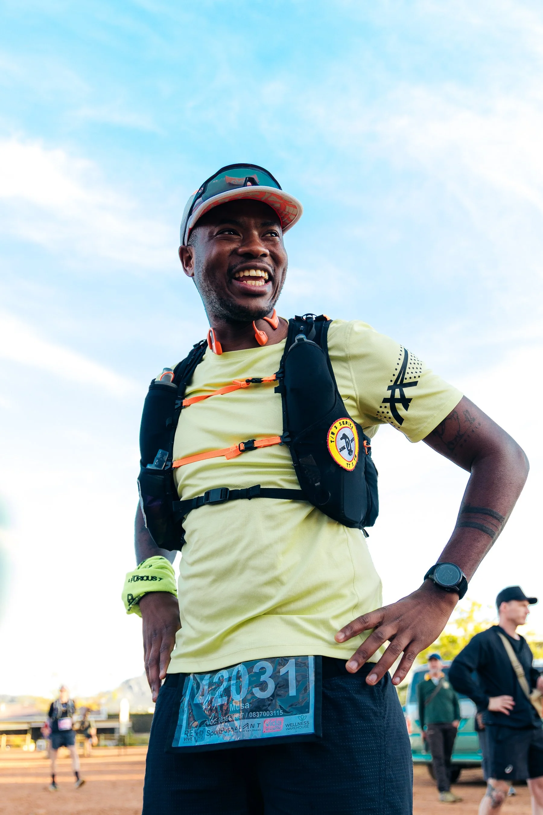 A man in a yellow running shirt and black running shorts, with a race bib number 2031, standing outdoors during a race event with other people in the background.