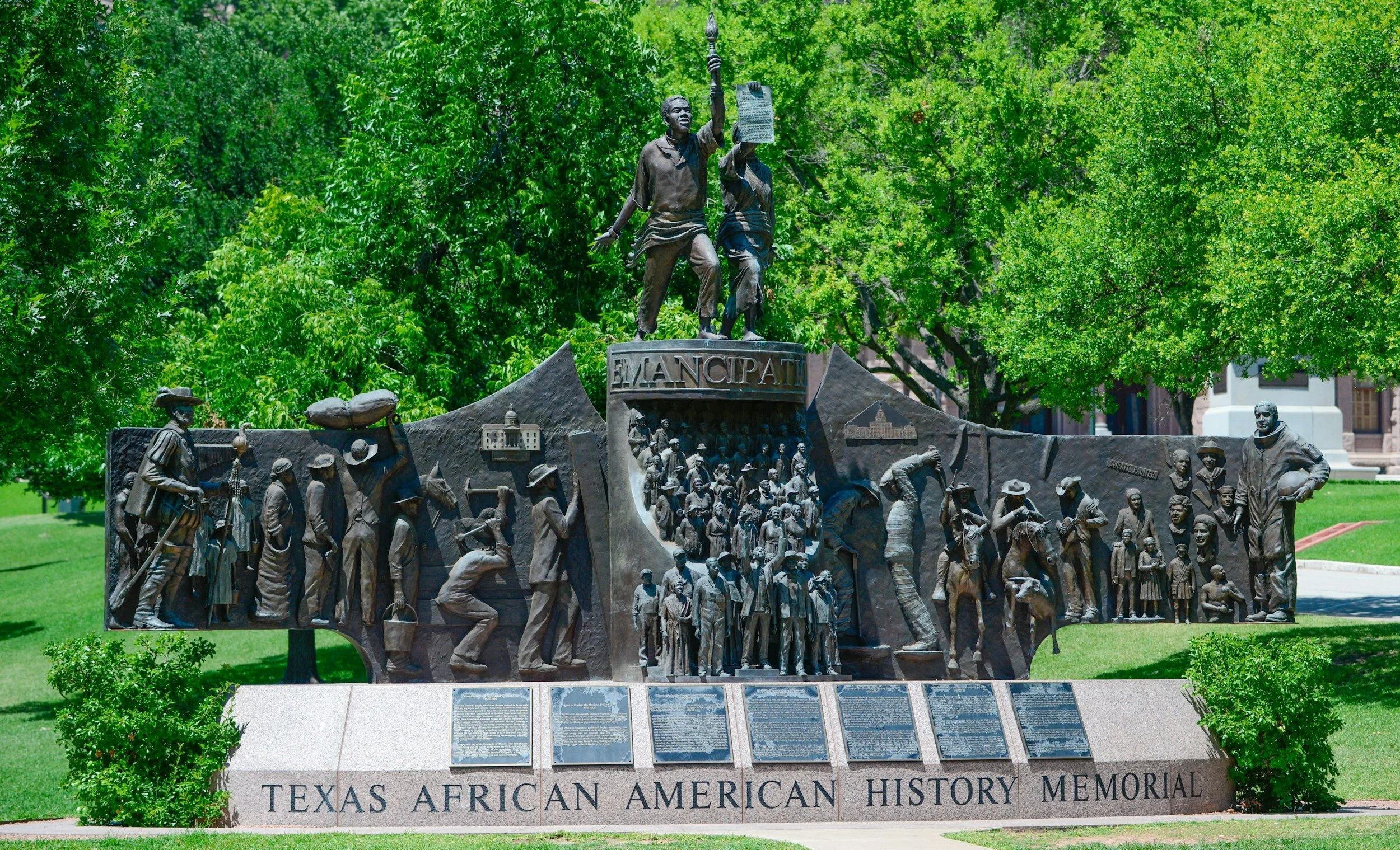 Texas African American History Memorial sculpture in a park setting, depicting historical figures and scenes, with 'Emancipation' inscribed prominently. Surrounded by green trees and grass.