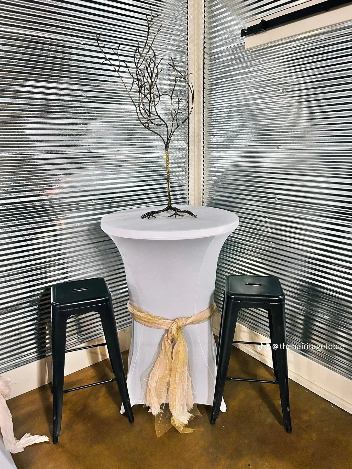 A tall, white high-top table with a tied gold cloth around its middle, flanked by two black stools. On top of the table is a decorative wire sculpture resembling a tree, placed in front of a corrugated metal wall.