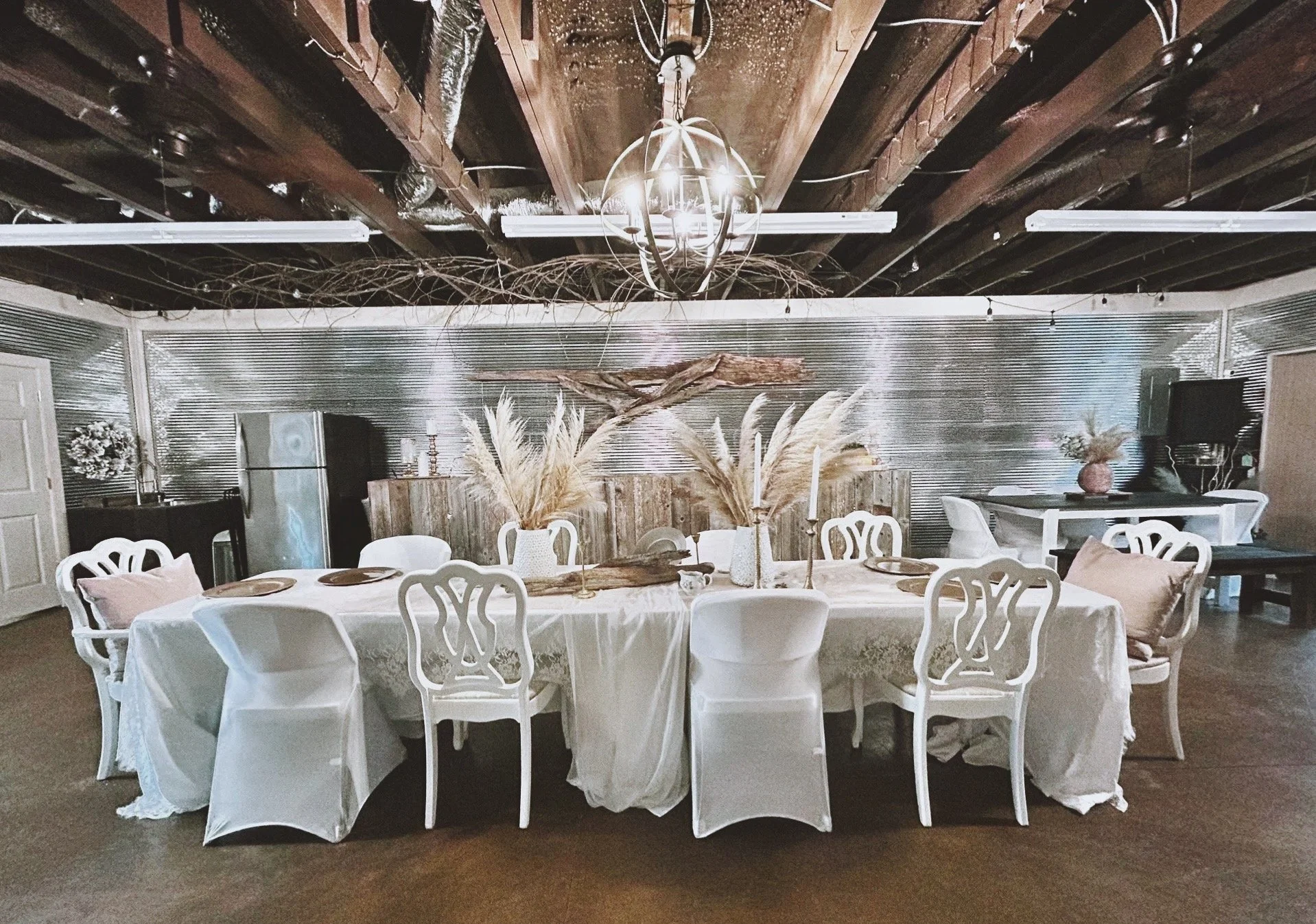 A round dining table with a white tablecloth, surrounded by white chairs, decorated with vases of dried pampas grass, candles, and gold plates, set in a rustic, elegant interior with wood and metallic decor.