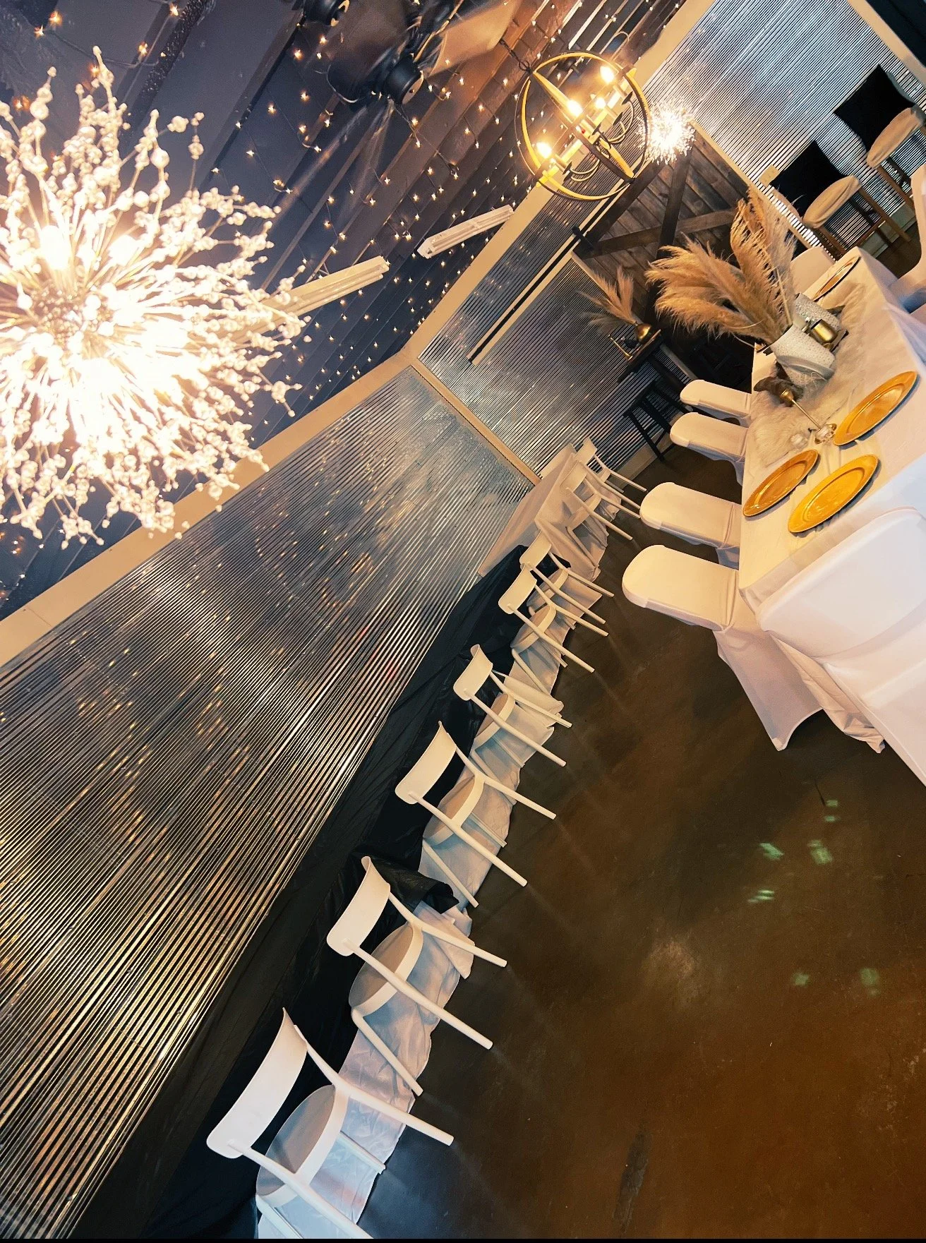 Elegant event setup with a long table covered in white tablecloths, gold plates, and decorative centerpieces featuring pampas grass and candles. There are white chairs arranged along the table, a backdrop with string lights and a festive chandelier, and a modern lighting fixture hanging from the ceiling. The setting appears to be indoors, with a sophisticated and warm atmosphere.