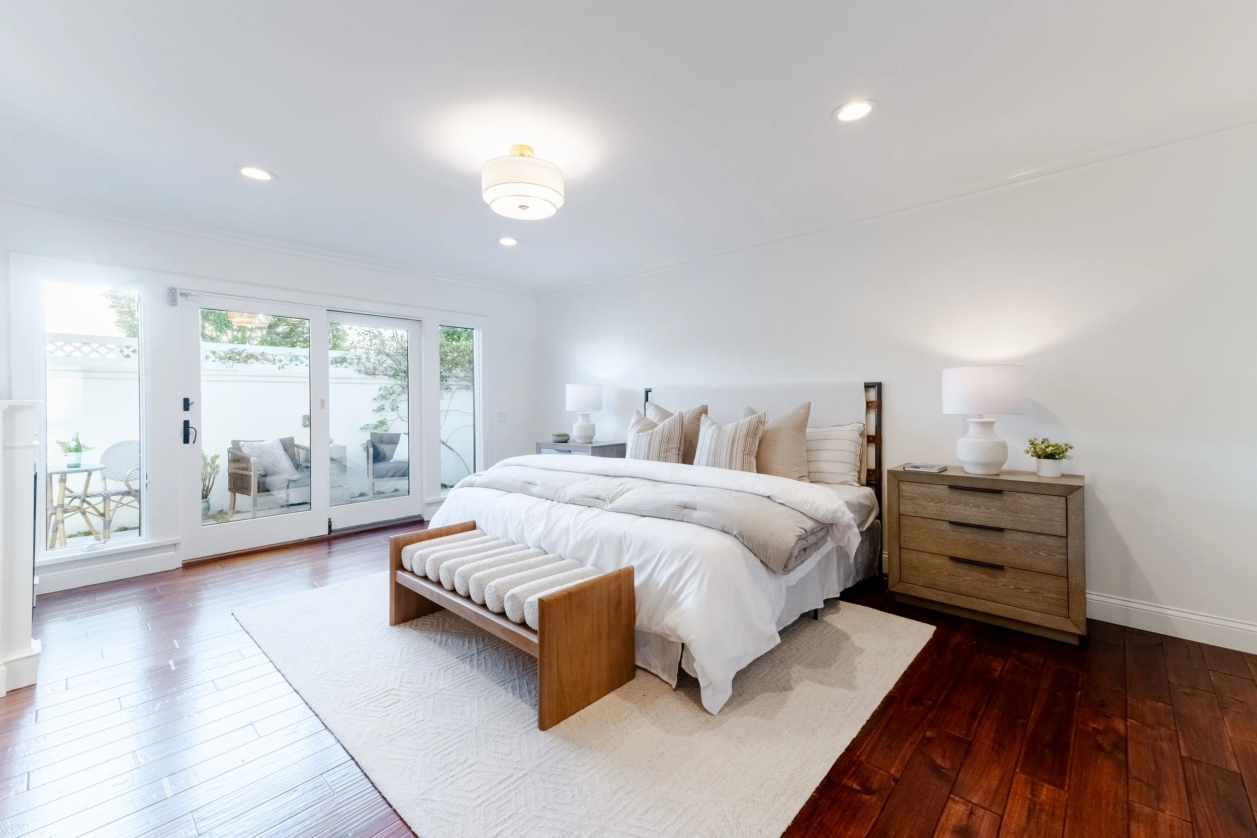 A bright bedroom with white walls and large sliding glass doors leading to a patio area. The bed has white and beige bedding, with multiple pillows. There are two wooden nightstands with lamps, and a wooden bench at the foot of the bed. The room has