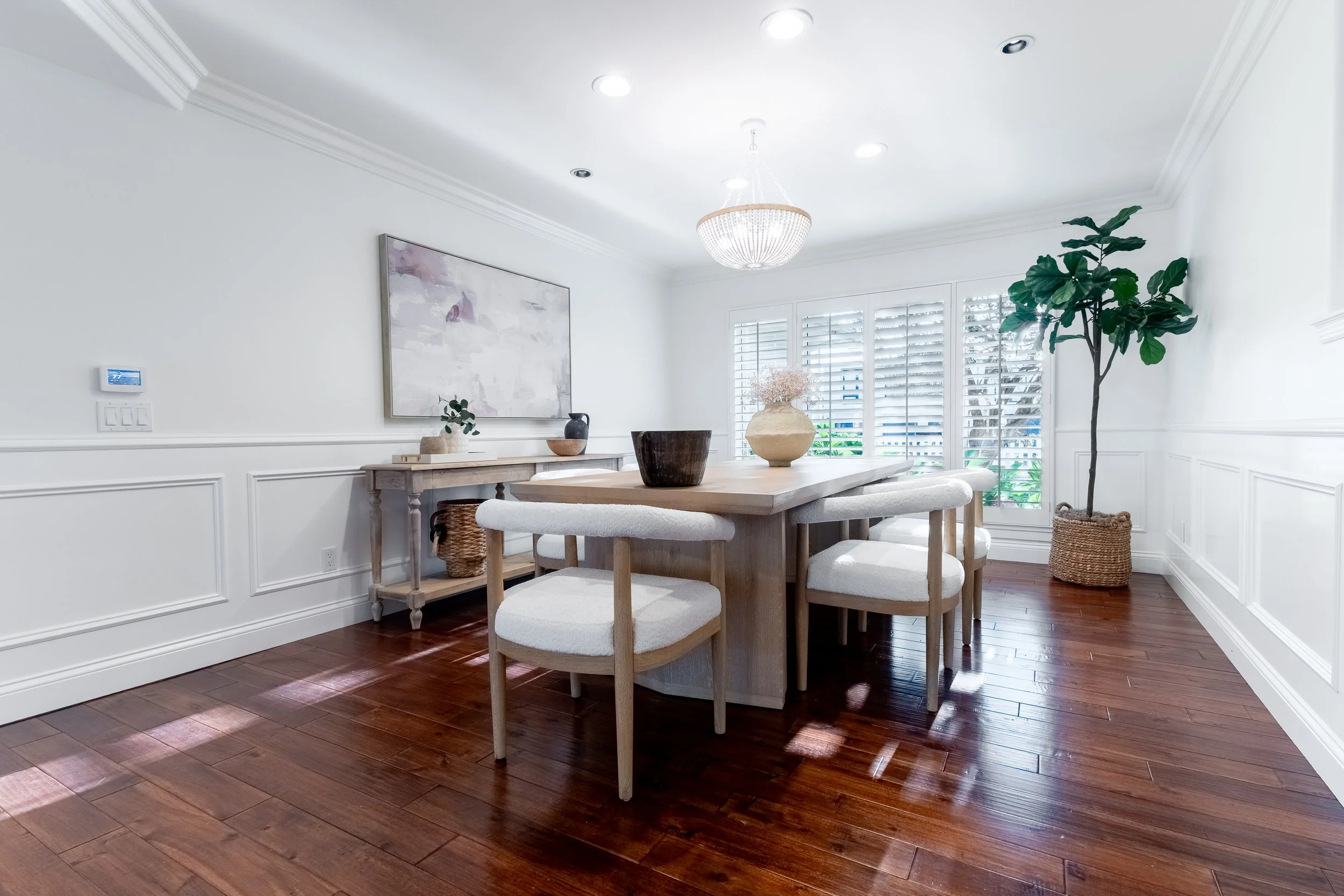A bright dining room with white walls, hardwood floor, a wooden dining table with six cushioned chairs, a large window with plantation shutters, a chandelier, potted plant, and artwork on the wall.