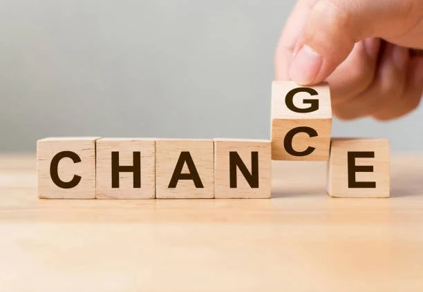 Wooden blocks spelling "CHANGE" with a hand flipping the "G" block to read "CHANCE" on a wooden surface.