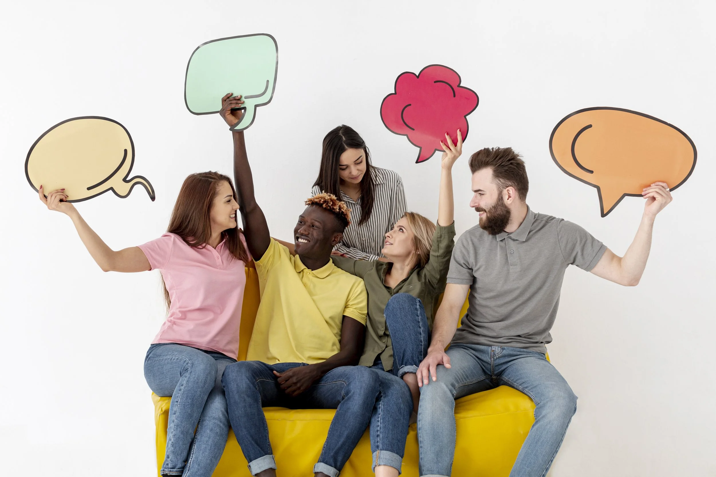 Group of five diverse people sitting on a yellow couch, holding colorful speech bubble signs, smiling and interacting.