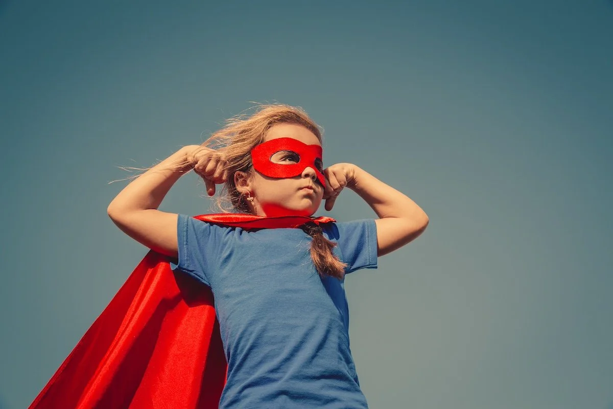 A young girl in a blue t-shirt, red cape, and a red mask flexes her biceps in a superhero pose.