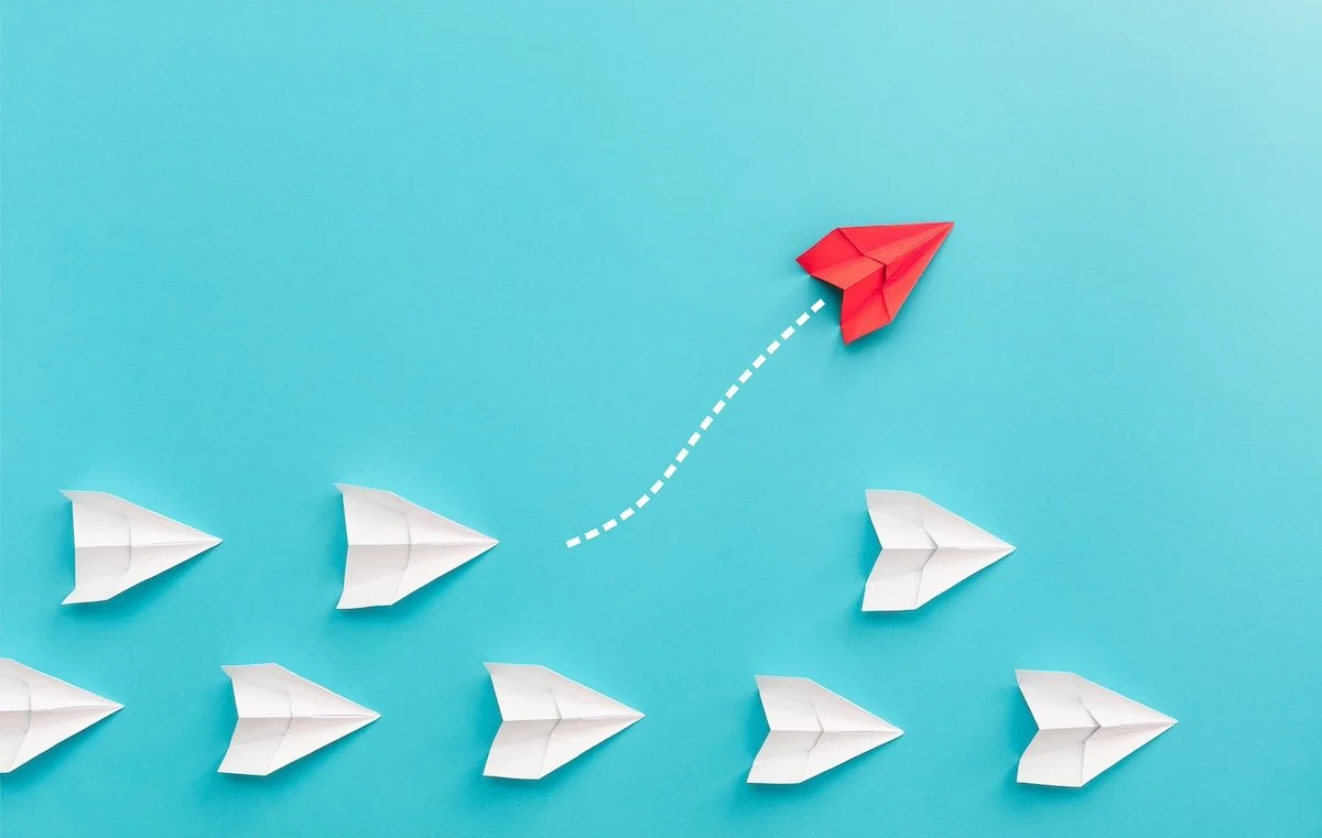 Rows of white paper airplanes, with one red paper airplane leaving the formation.