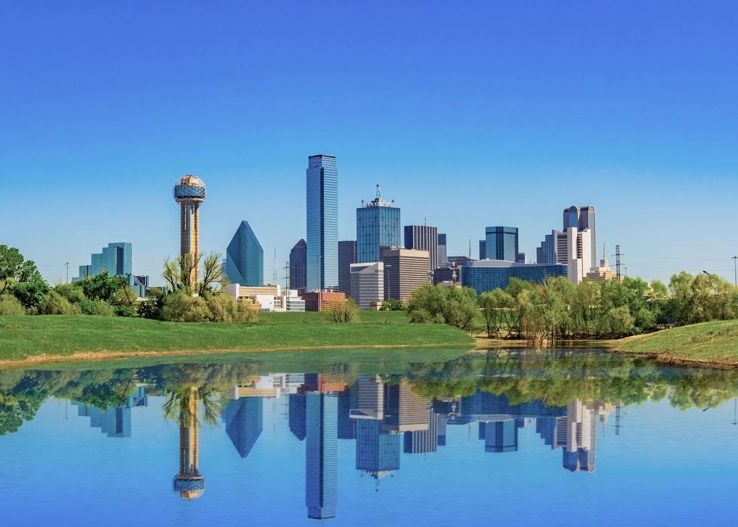Dallas skyline with modern skyscrapers and a reflection in a calm body of water with greenery in the foreground.