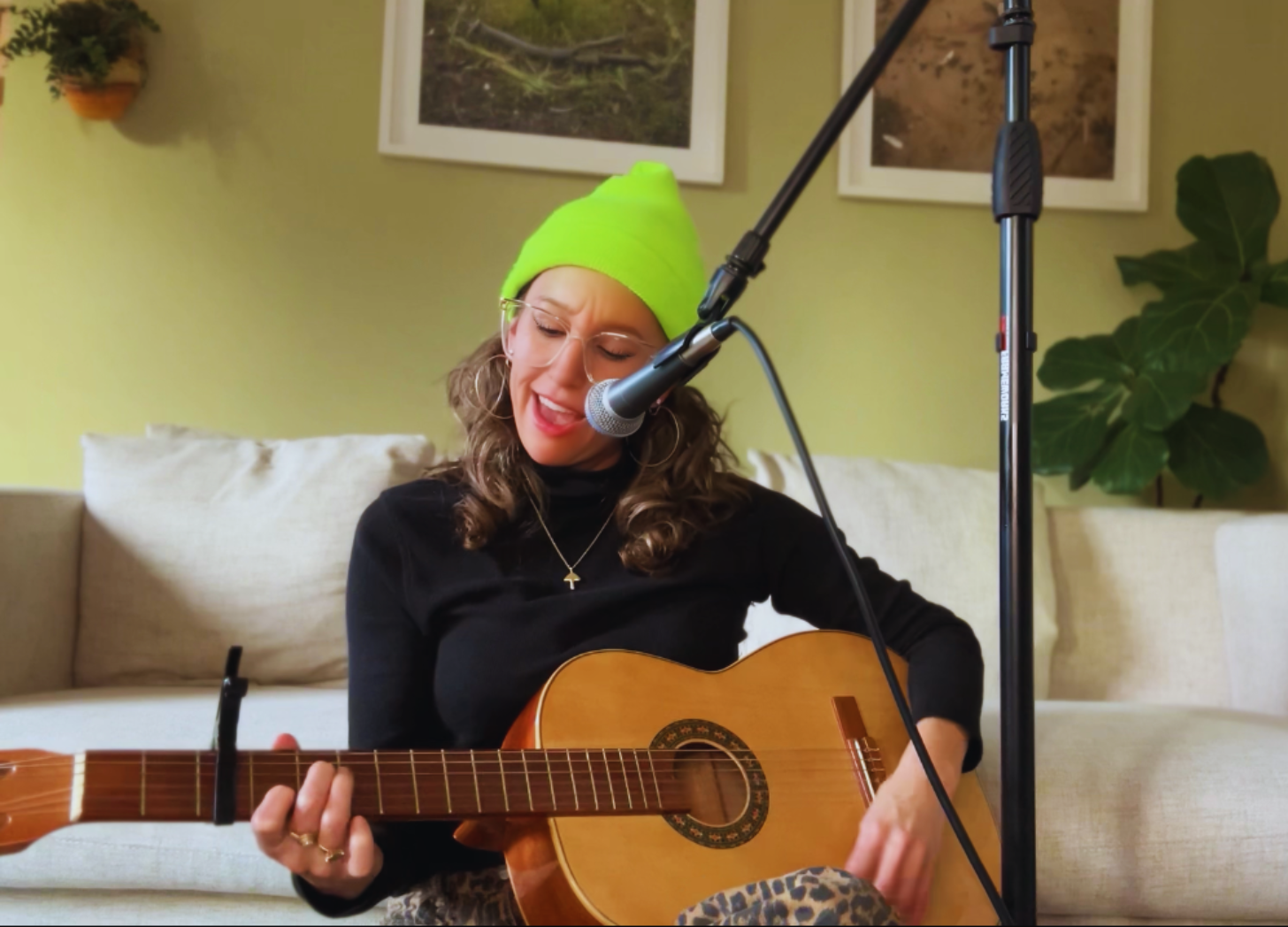 Person wearing a lime green beanie and glasses, playing acoustic guitar and singing into a microphone, sitting in a cozy living room with framed pictures on the wall.