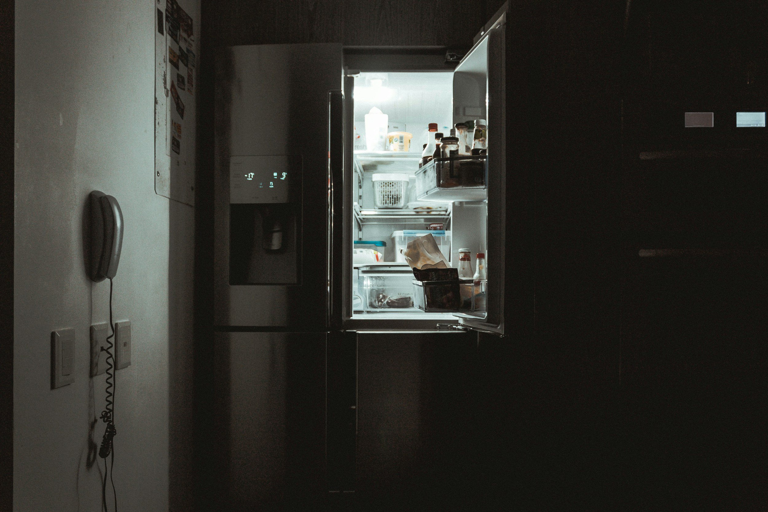 Modern kitchen with stainless steel refrigerator, microwave, stove, and black backsplash, white cabinets, dark wooden floor, and two black pendant lights hanging above a white kitchen island with two black stools.