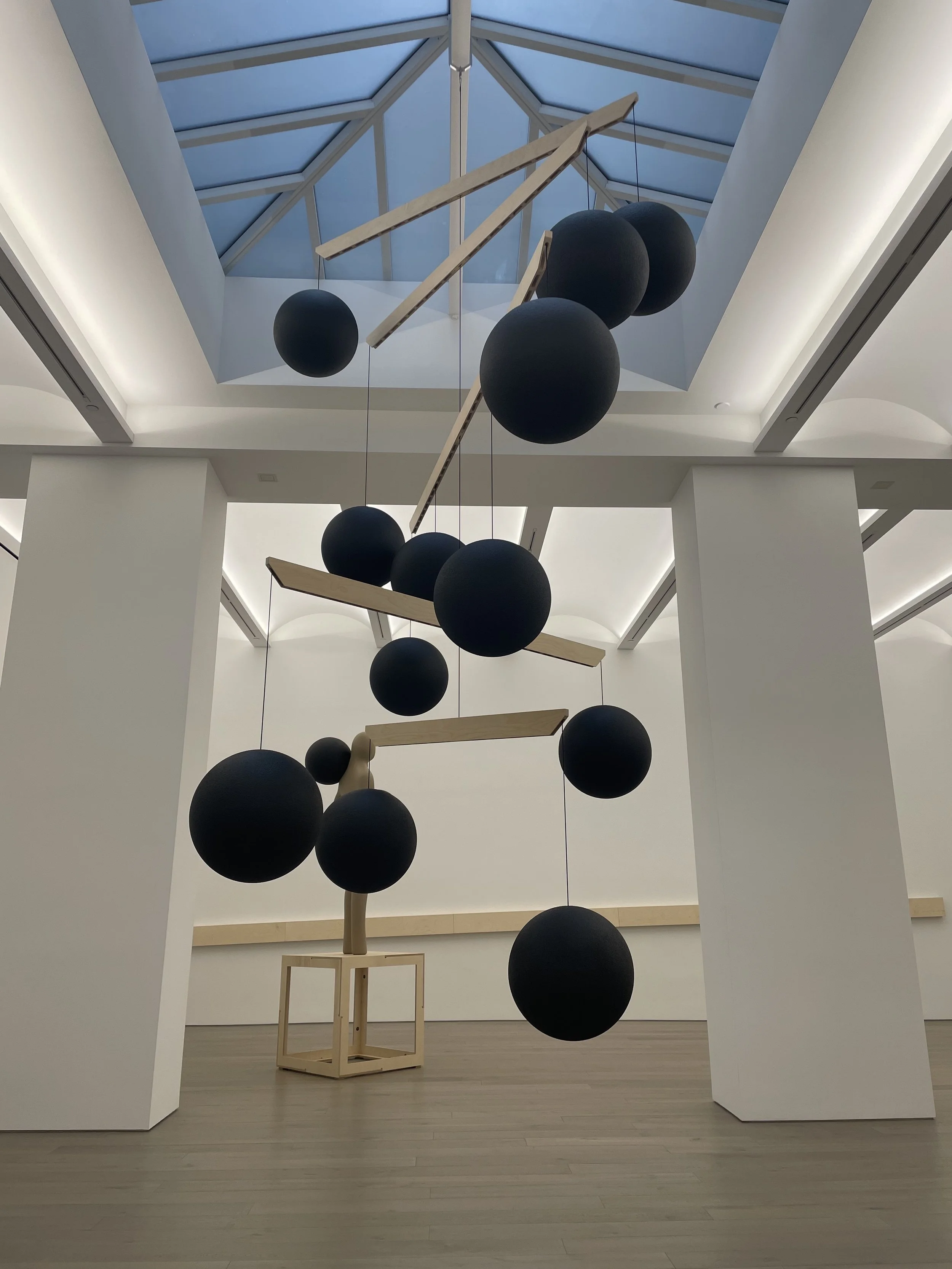 Modern art installation featuring black spherical shapes hanging from wooden beams inside a bright, spacious gallery with a skylight.