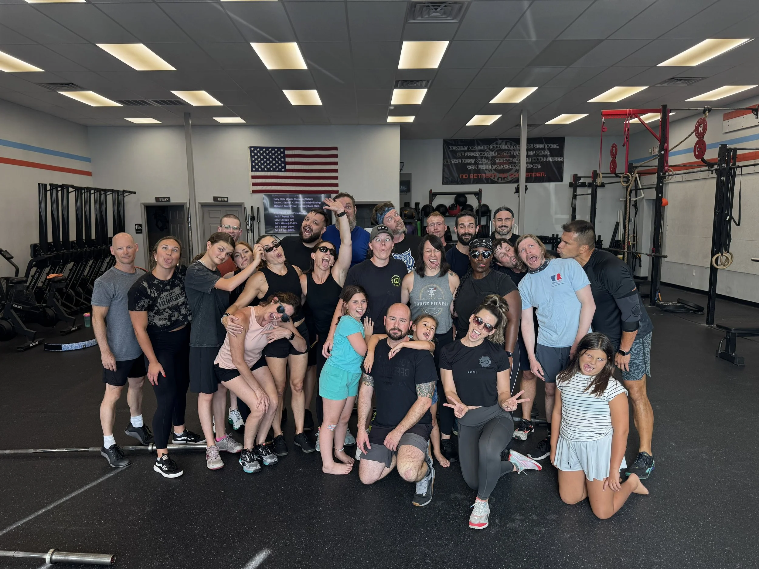 Group of people at a gym, smiling and posing for a photo with gym equipment and an American flag in the background.
