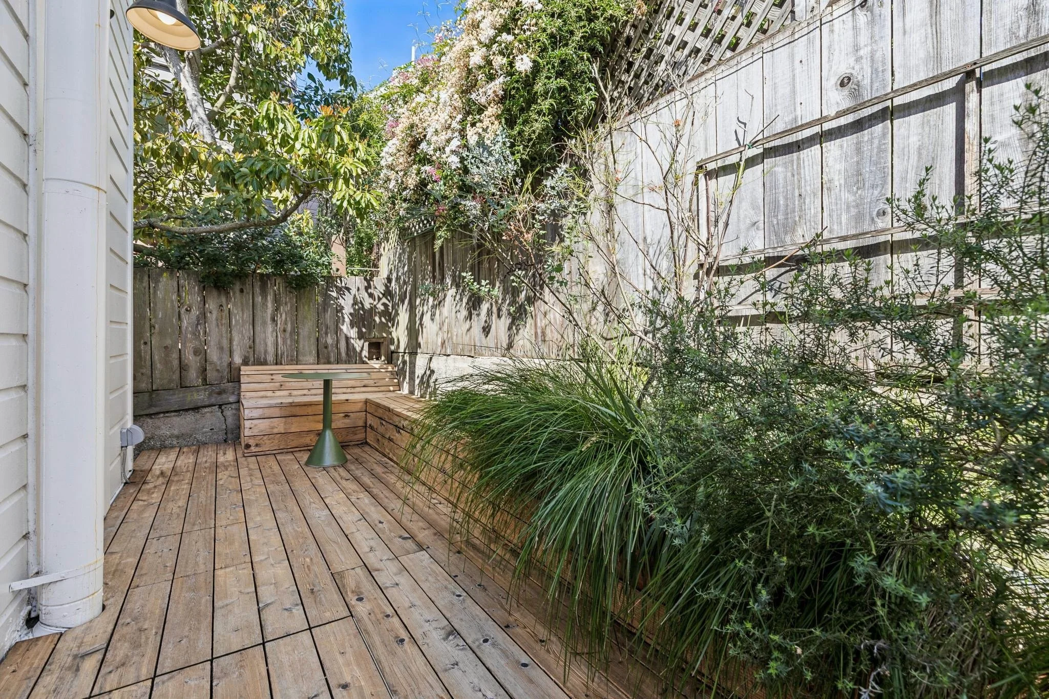 Empty wooden outdoor patio with built-in bench, small round table, green plants, and a weathered wooden fence with flowering vine on top