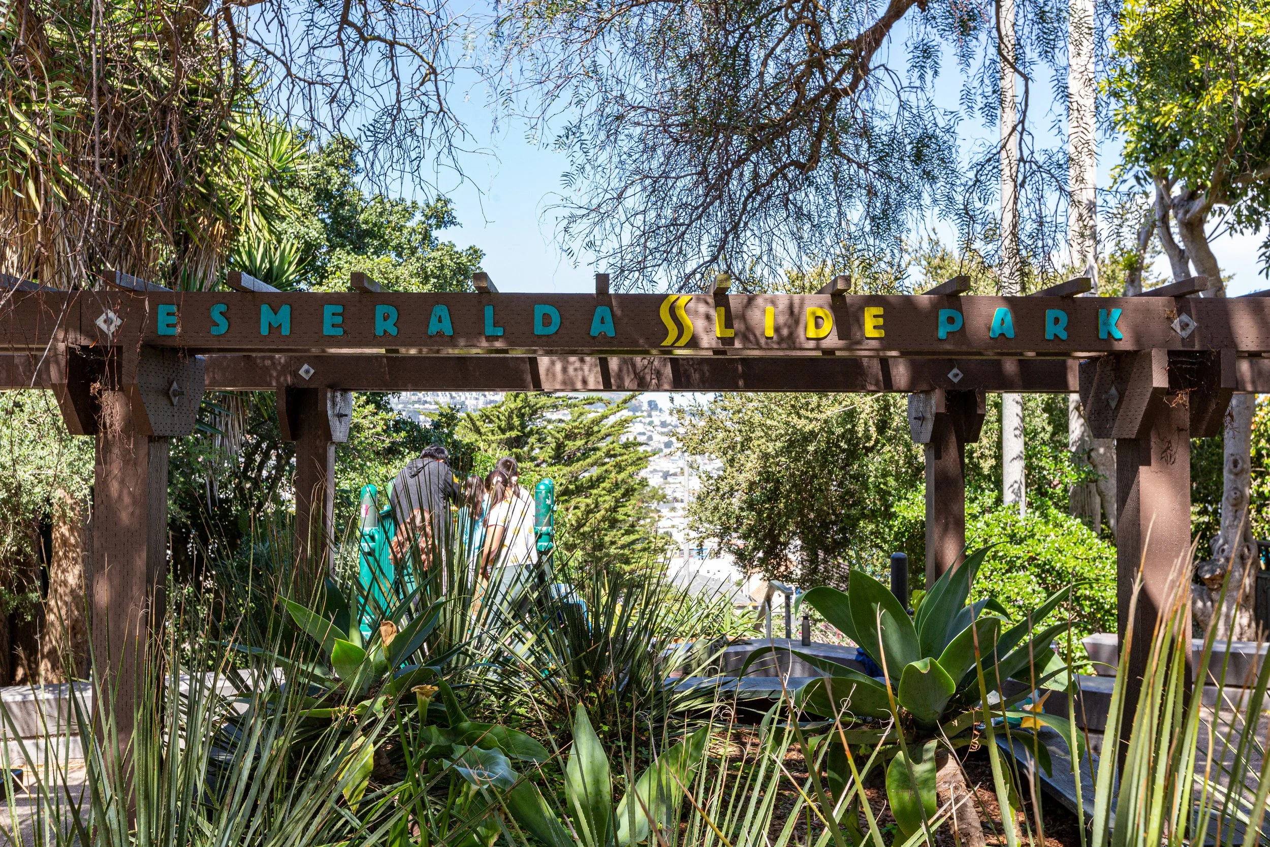 Entrance sign for Esmeralda Slide Park with people walking through lush greenery and trees in the background.