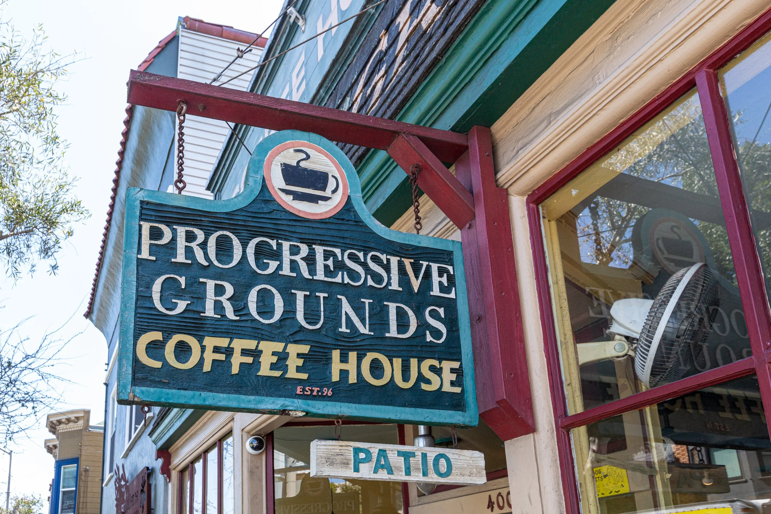 Sign for Progressive Grounds Coffee House with a patio menu below, hanging outside a colorful building with large windows and a ceiling fan visible inside.