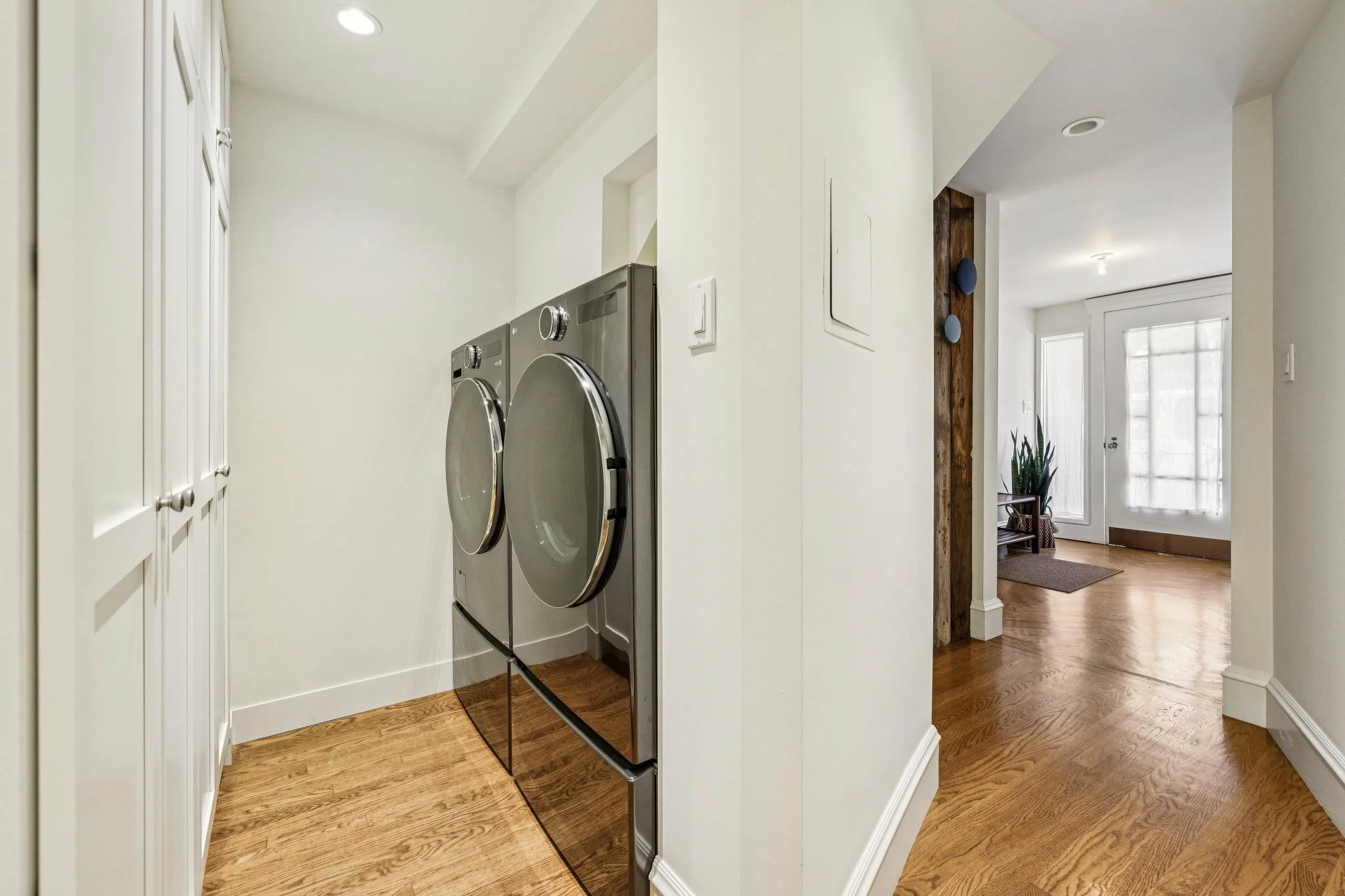 Modern laundry room with washer and dryer, white cabinets, wooden floor, and doorway leading to a bright living space.