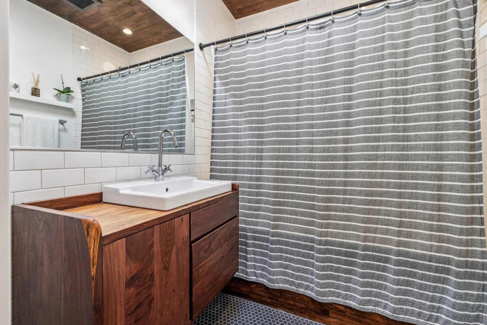 Bathroom with wooden vanity, white sink, large mirror, white tile walls, gray striped shower curtain, small black hexagon floor tiles, and a white towel on a silver towel rack.
