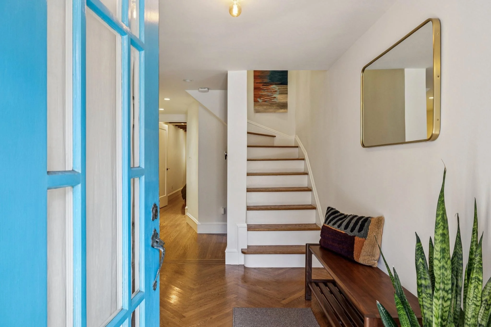 Entrance hallway with a blue front door, wooden flooring, a staircase with white risers and wooden treads, a mirror with a gold frame on the wall, a wooden bench with a cushion, a potted plant, and a piece of artwork at the top of the stairs.