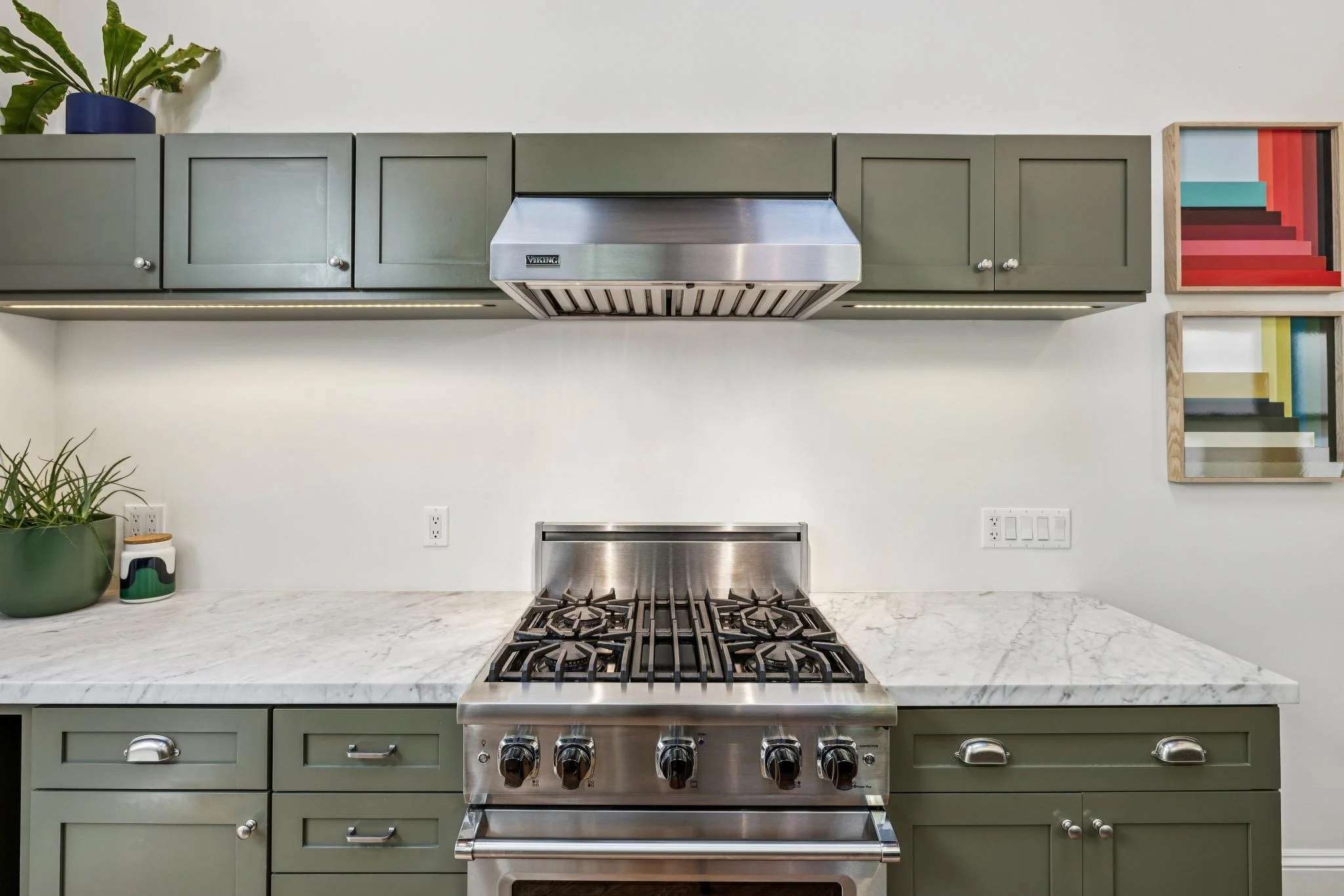 Modern kitchen with green cabinetry, stainless steel stove and range hood, white marble countertop, potted plant, and colorful geometric wall art.