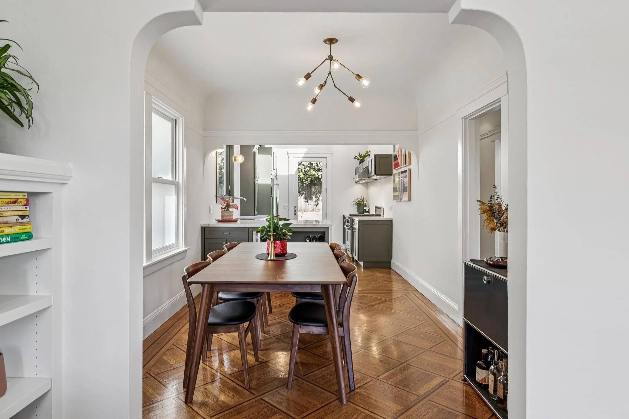 Dining room with wooden table and black chairs, gray kitchen cabinets, hardwood floors, and white walls with large windows.