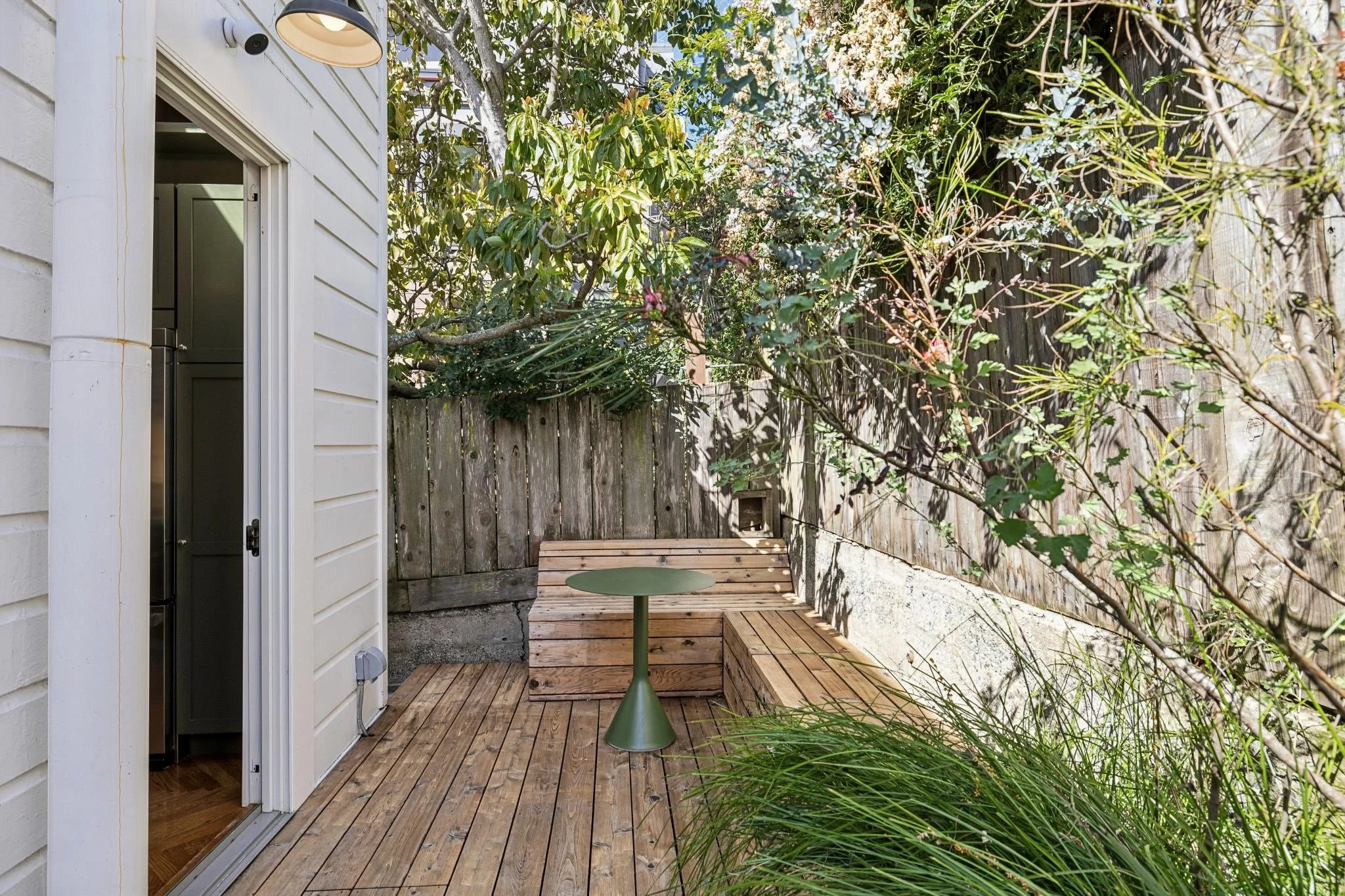 Small backyard patio with wooden decking, a built-in wooden bench, a round green table, and surrounding greenery, including a tree, bushes, and grass, with a wooden fence in the background.