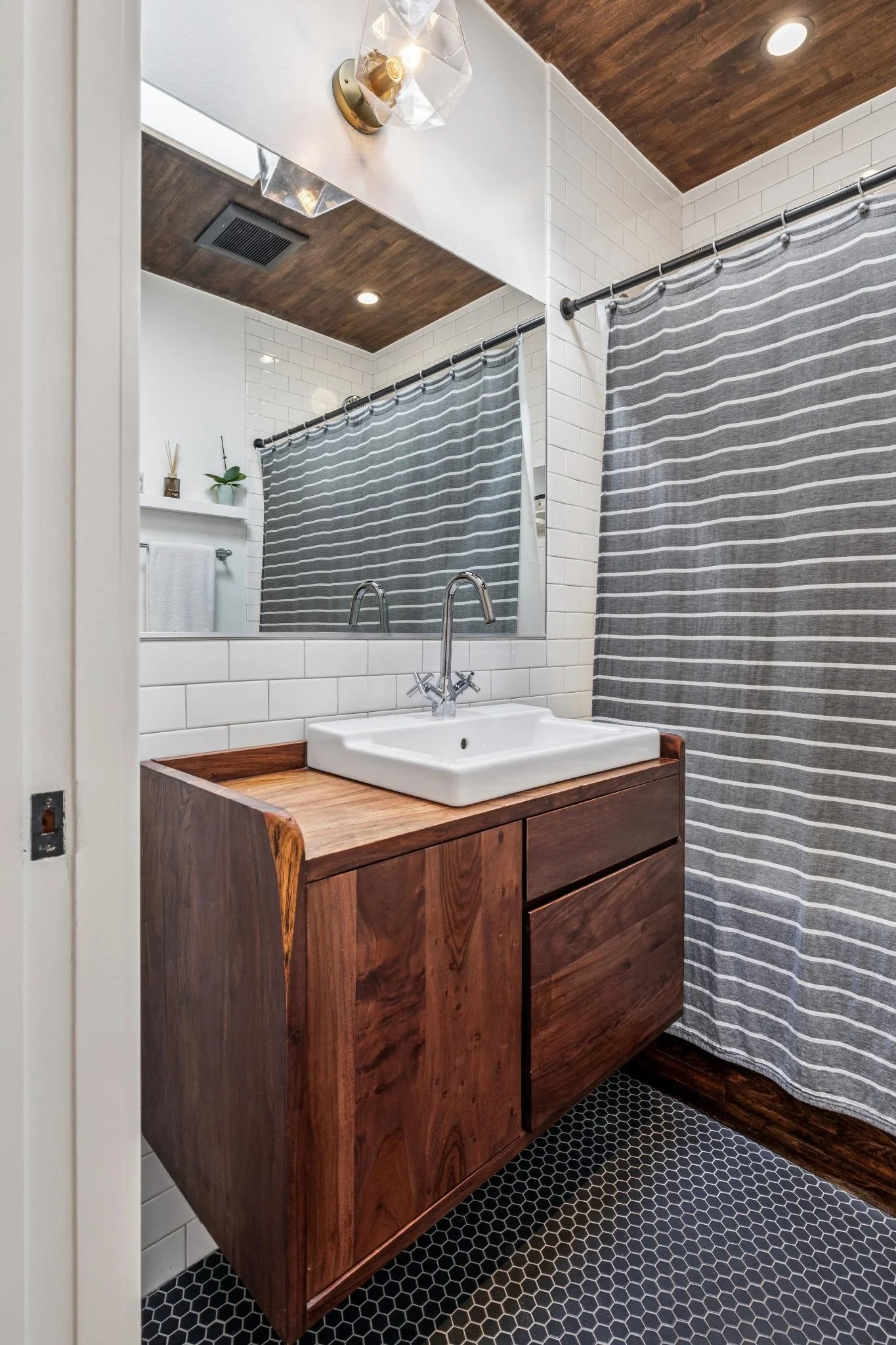 Bathroom with a wooden vanity, white vessel sink, striped gray shower curtain, white tiled wall, round black tile floor, and a large mirror.