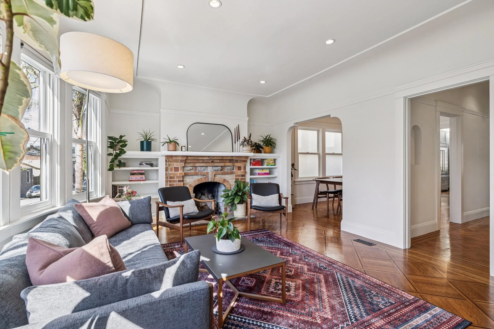 Bright living room with large windows, gray sofa with pink pillows, black armchairs, a brick fireplace with a mirror above, wooden floor, and assorted houseplants.