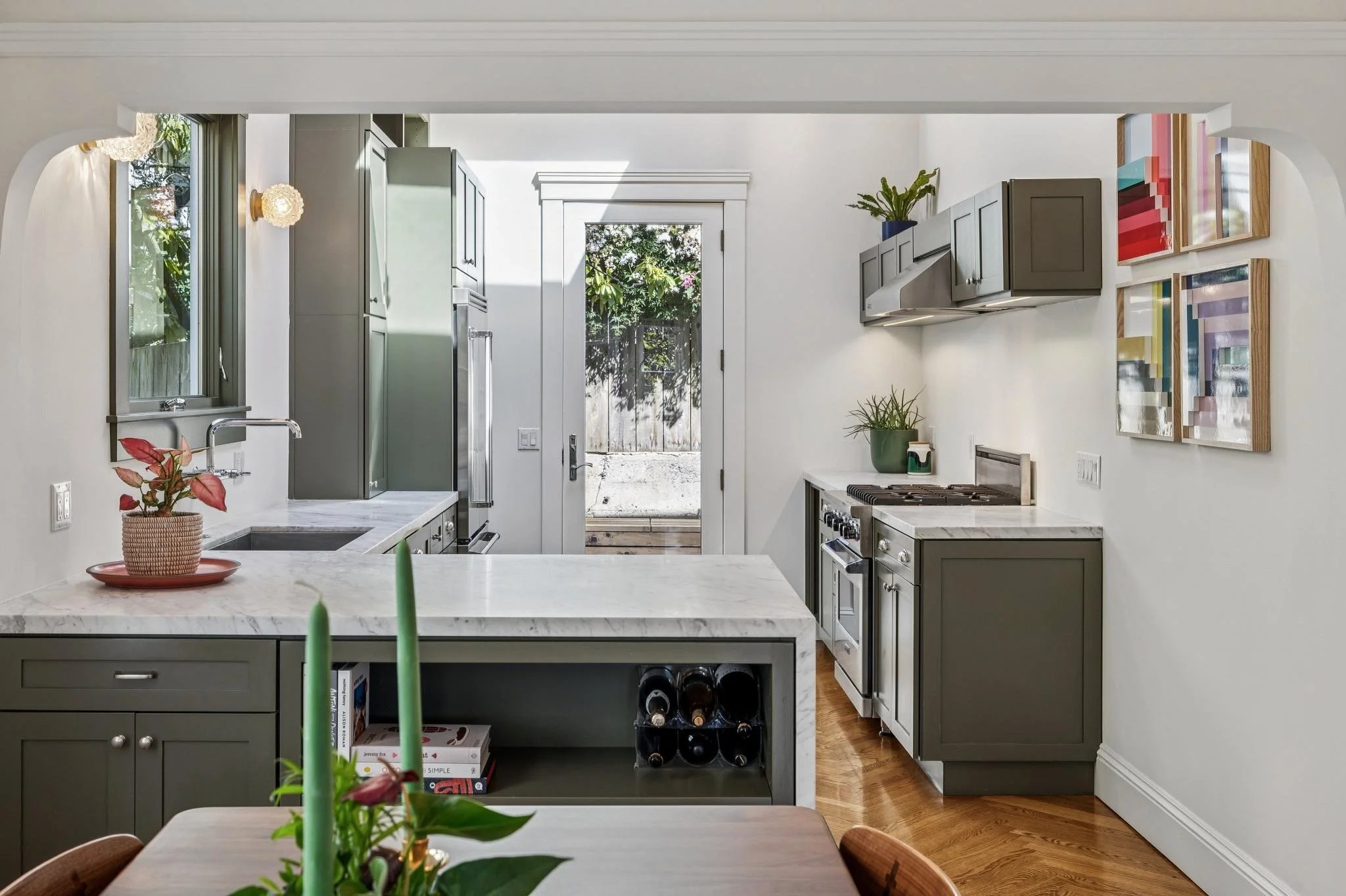 Modern kitchen with gray cabinets and white marble countertops, a small wine storage area, a window and a glass door leading outside, and colorful artwork on the wall.