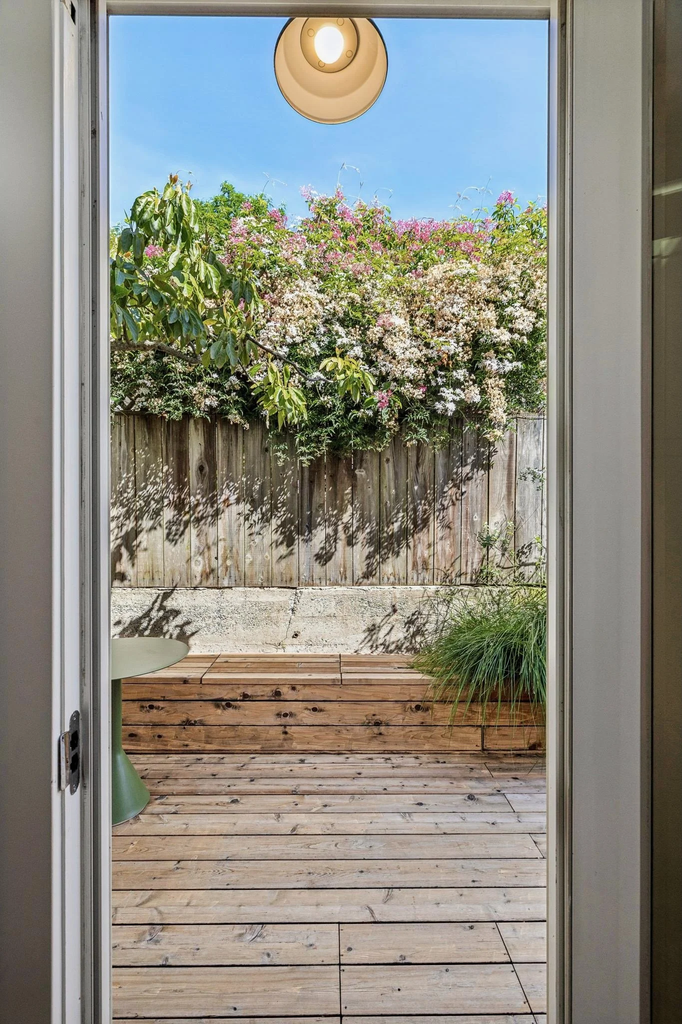 View through a doorway of a wooden deck, a weathered wooden fence, flowering bushes, and a clear blue sky.
