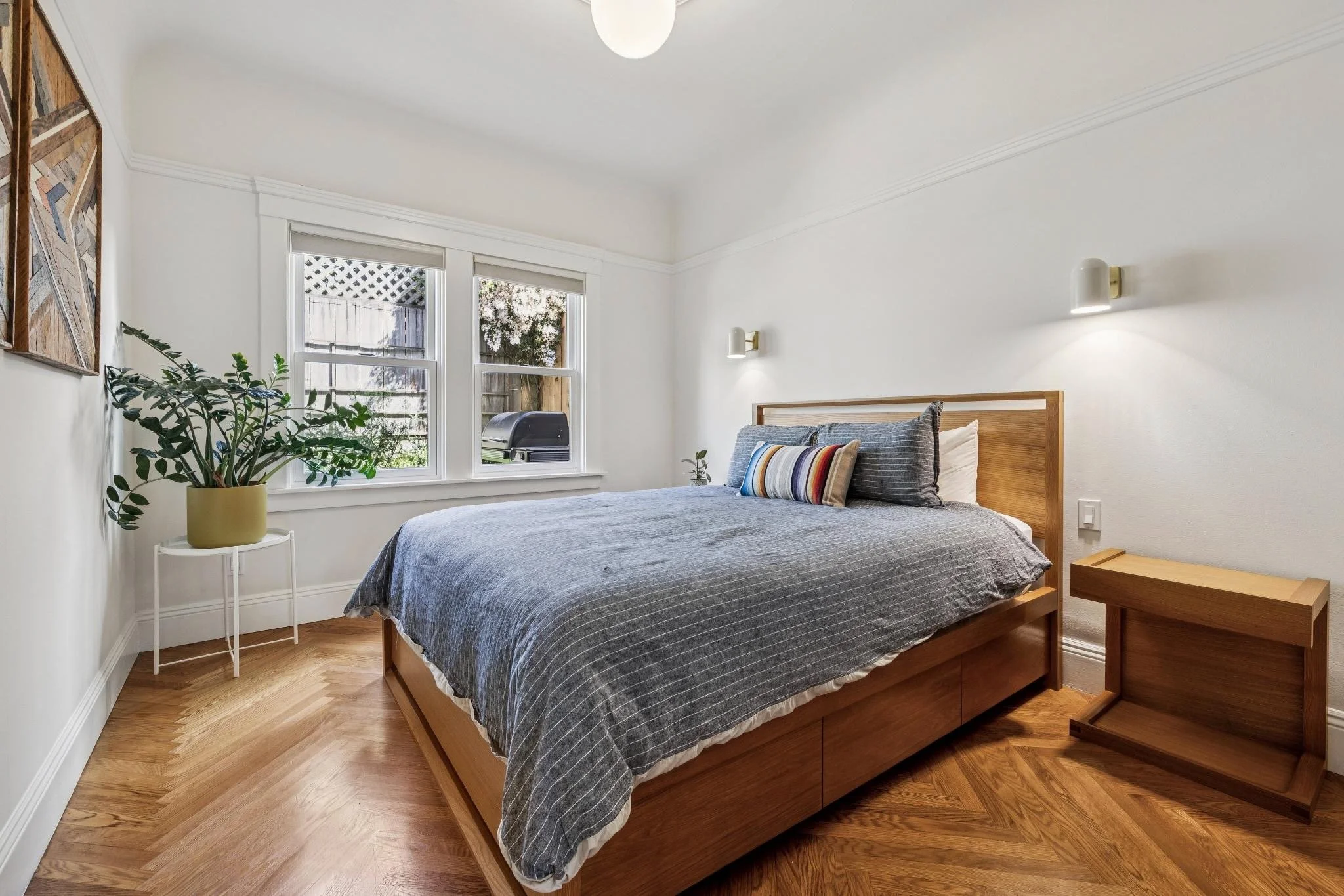A bedroom with a large bed, gray striped bedding, wood headboard, two wall-mounted lights, a small wooden nightstand, and a plant in a yellow pot by the window.