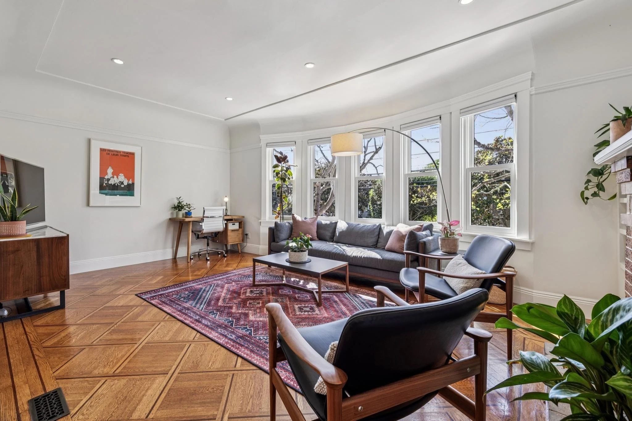 Bright living room with hardwood floors, a gray sofa with pink cushions, a black armchair, a wooden coffee table, a corner desk with a chair, and large windows with trees outside.