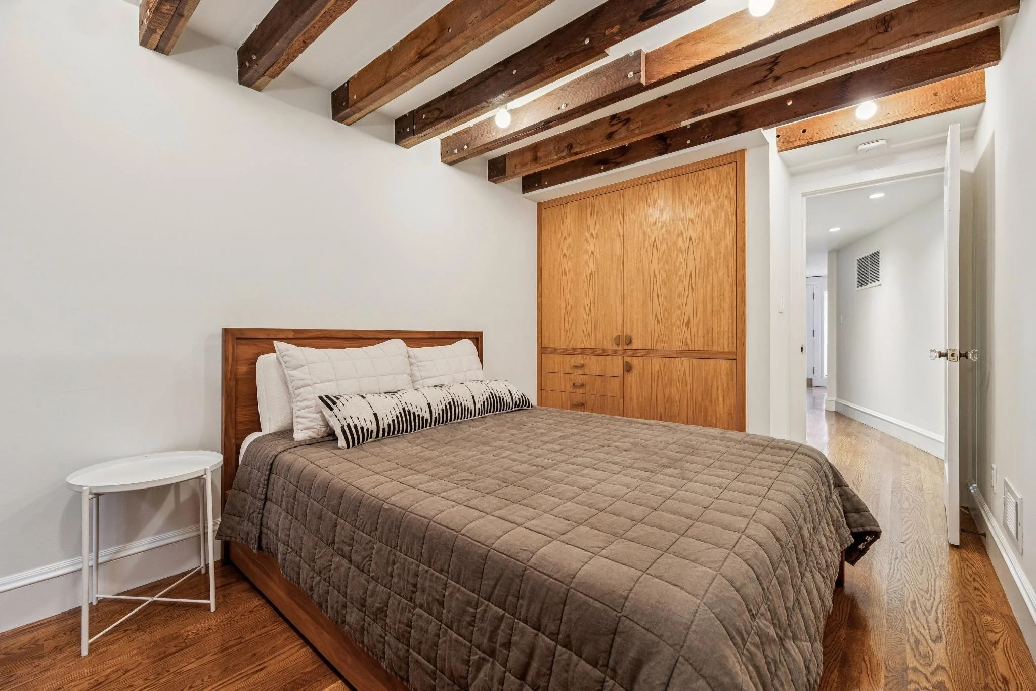Comfortable bedroom with a wooden bed frame, beige quilted bedspread, decorative pillows, a white side table, in a room with wooden beams on the ceiling and a built-in wooden closet.