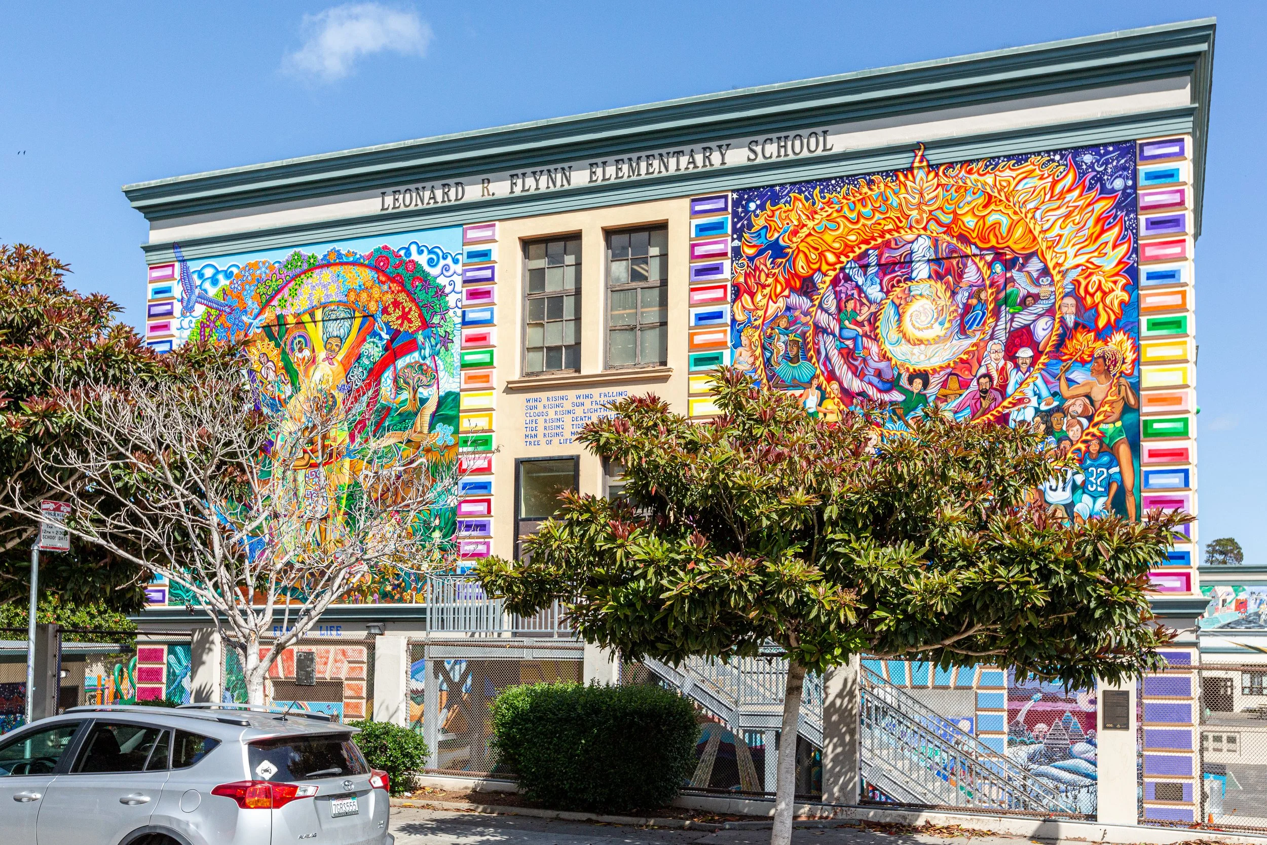 Colorful mural paintings on the exterior wall of Leonard R. Flynn Elementary School, featuring vibrant scenes of nature, people, and flames, with the school's name displayed at the top. There are trees and a parked car in the foreground.