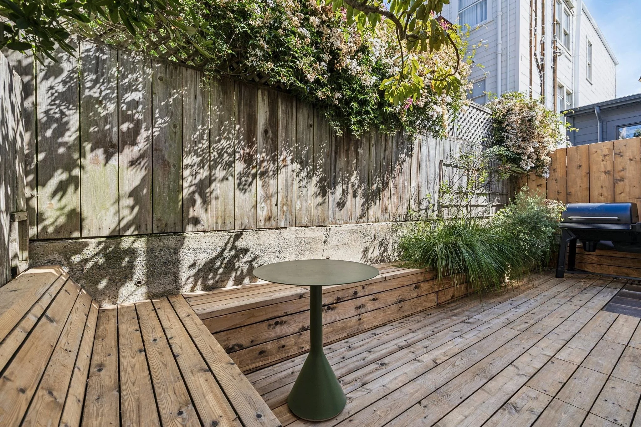 A backyard wooden deck with a small round table, a bench, a grill, and lush green plants with flowers along a weathered wooden fence, shaded by tree branches.