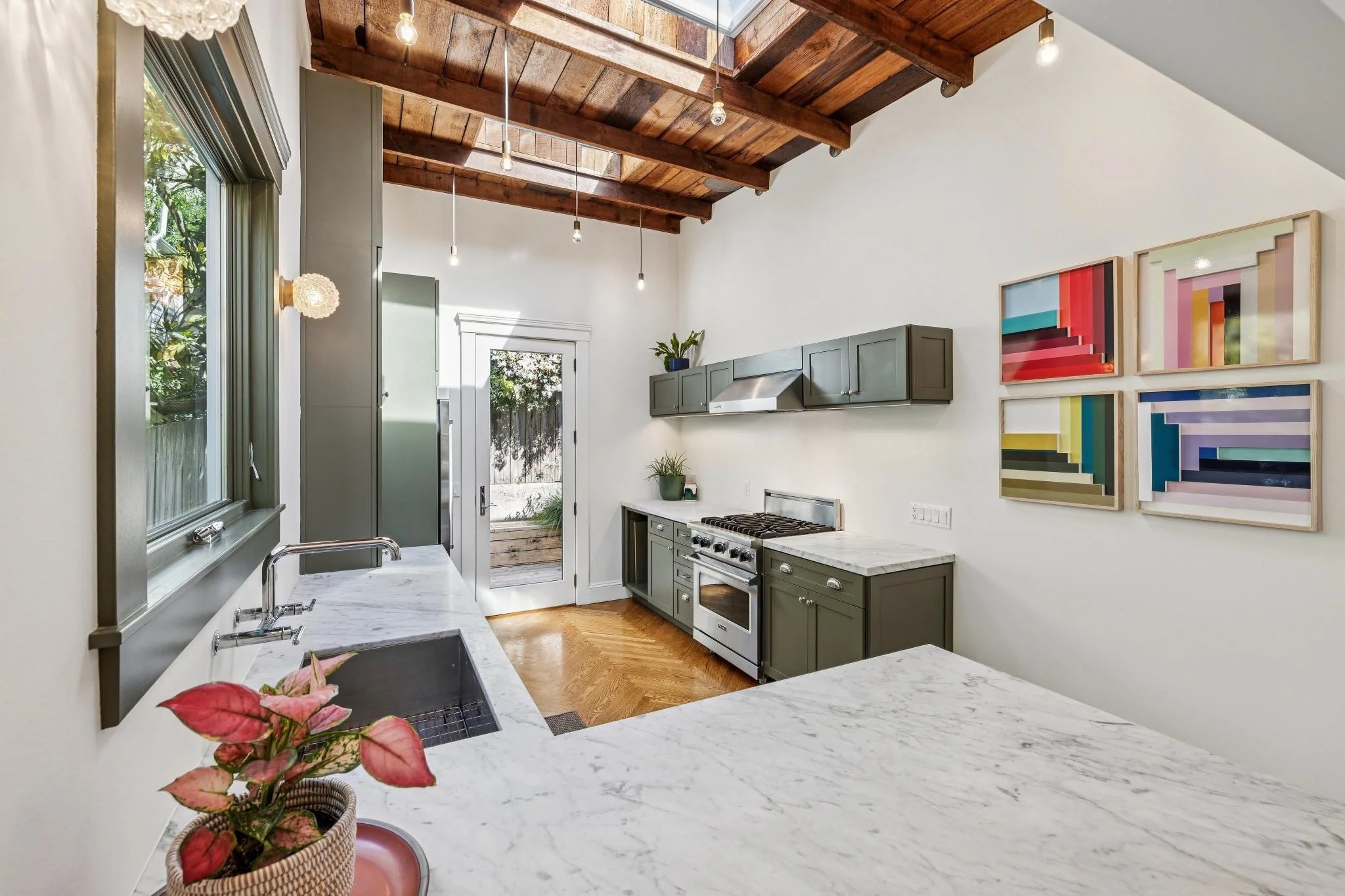 Modern kitchen with green cabinets, marble countertops, wooden ceiling beams, wall art, and a door leading outside.
