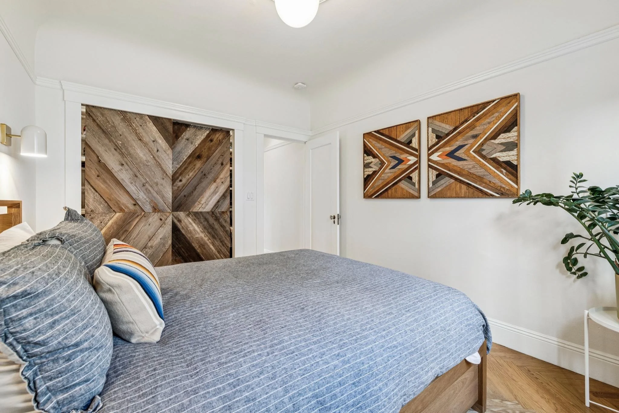 A bedroom with a bed covered in a gray striped bedspread and several pillows, including one with multicolored stripes. On the wall above the bed are two large, abstract wood art pieces. To the left is a white wall with a small modern light fixture. A