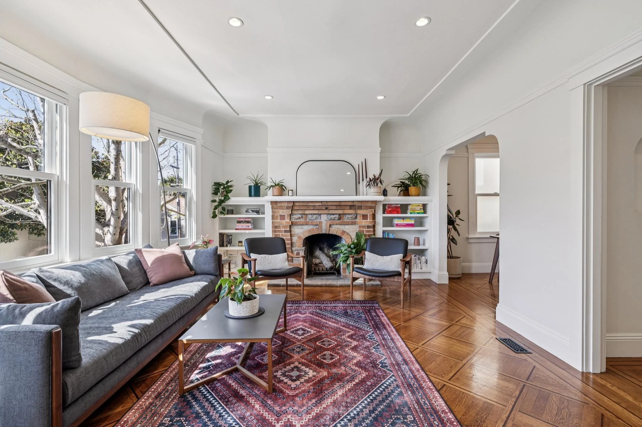 Living room with a gray sofa, a Persian rug, a brick fireplace with a mirror above, built-in shelves with books and plants, and three black chairs facing the fireplace.